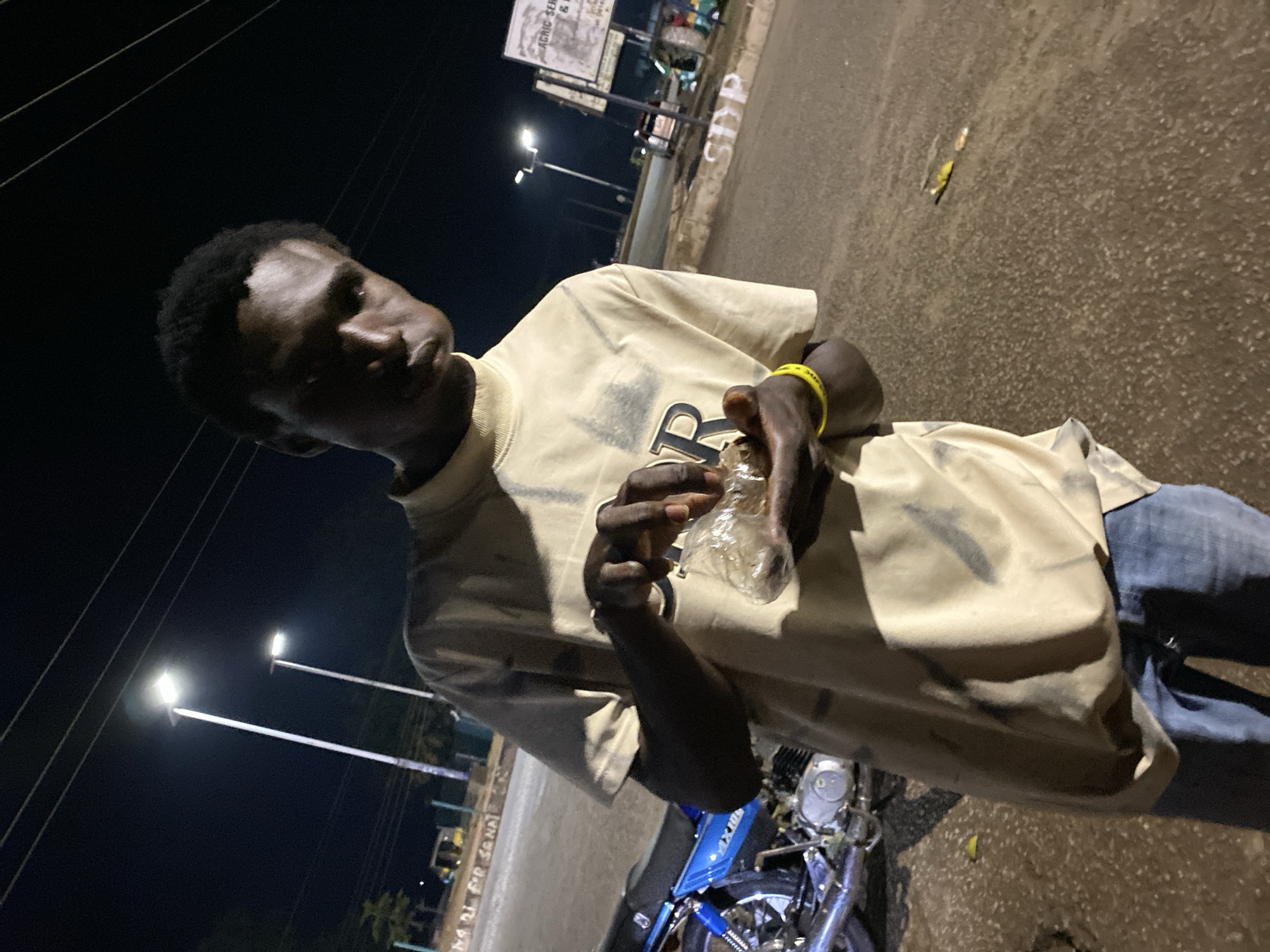 A young Black male, appearing to be in his late teens or early twenties, stands outdoors at night in what appears to be an urban environment. He is wearing a light-colored, long-sleeved t-shirt with a graphic design on the front, and blue jeans. He is holding a clear plastic bag in his hands, and appears to be examining its contents. To his left, a blue motorcycle is parked. The background shows streetlights illuminating the area, and power lines are visible overhead. Further in the background, there are signs and some buildings. The ground is paved and appears to be a road or sidewalk. The scene suggests a moment of pause or observation, possibly in a commercial or residential area at night. The location is identified as Bukuru, Nigeria.