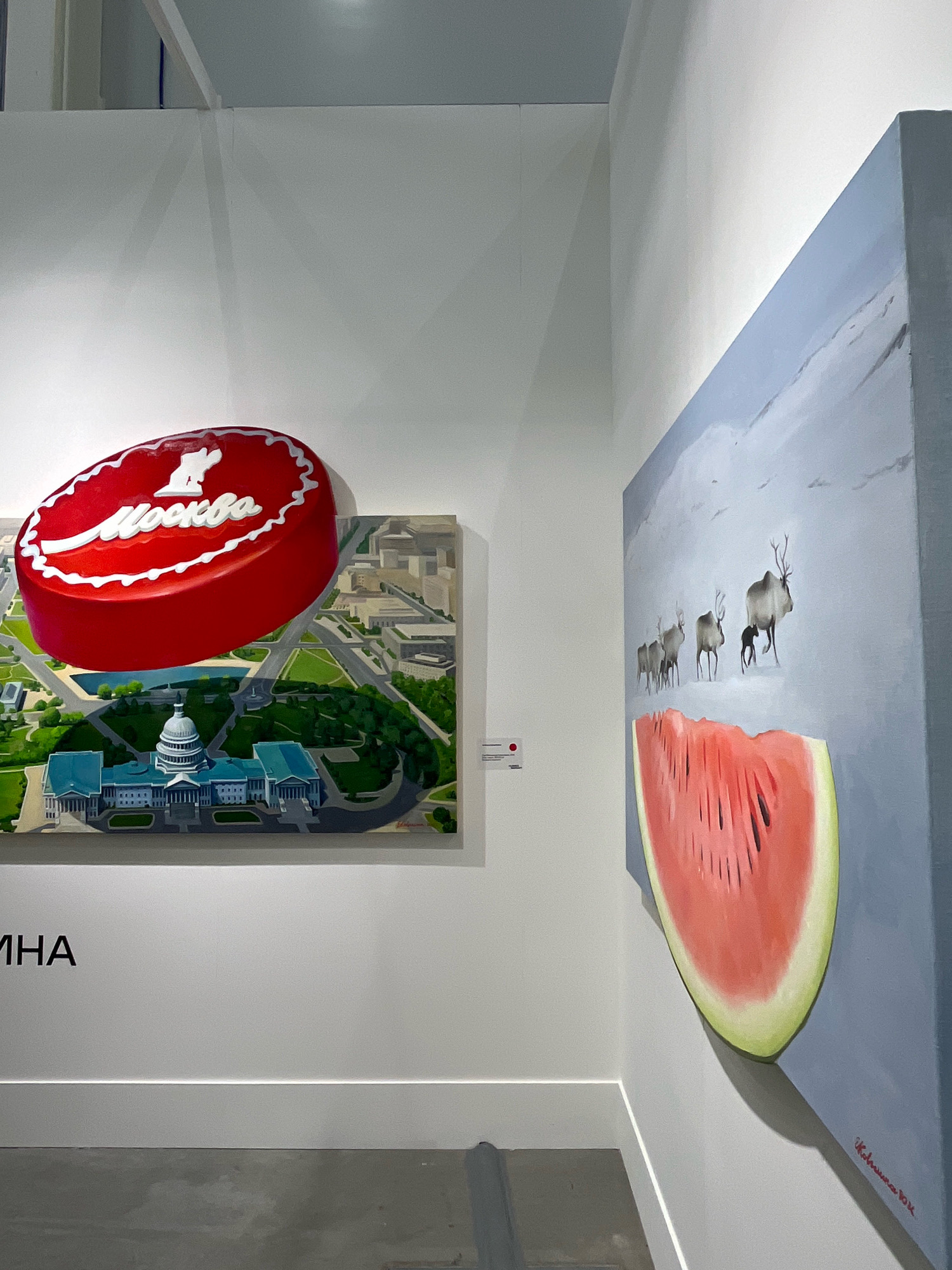 The image displays two paintings in what appears to be an art gallery. The painting on the left is a surrealist piece featuring a giant red candy with the word "Mockba" (Moscow) written in white script, hovering over an aerial view of Washington D.C., specifically the U.S. Capitol building and the surrounding National Mall. The painting on the right is a landscape depicting a herd of reindeer walking across a snowy plain, with a large slice of watermelon prominently featured in the foreground. The text "МНА" is visible on the wall below the left painting, likely an abbreviation related to the exhibition or venue. The location is confirmed to be in Khimki, Russia, indicated by the visible Cyrillic text. The environment is a clean, well-lit indoor space with white walls and a grey floor. There are no people visible in the image.