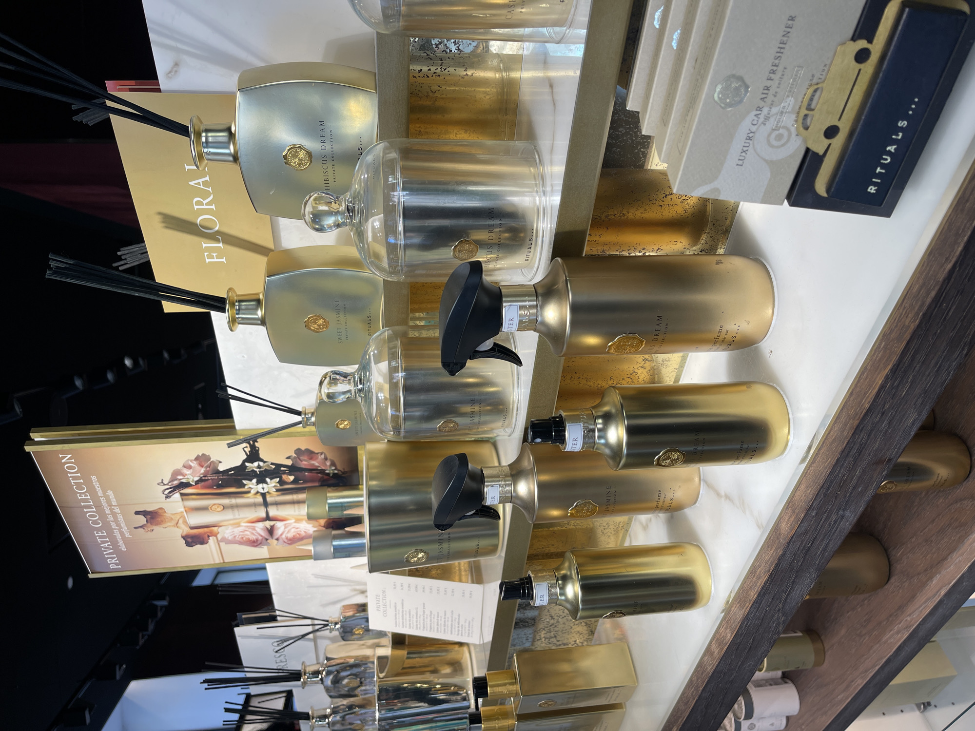 This image captures a well-lit retail display of the "Rituals" brand's "Private Collection" products, strongly suggesting a store located in Valencia, Spain, due to the prominent Spanish language on various labels (e.g., "elaboradas por los mejores maestros perfumistas del mundo," "Ambientador," "Velas perfumadas") and Euro pricing (€) on a visible price list.
The scene features multiple shelves constructed with a white, marble-patterned surface and dark wooden trim. The backdrop behind the main products is a textured, mottled gold or bronze finish.
The display showcases a variety of home fragrance items, predominantly in metallic gold and bronze-colored packaging with black accents and clear glass elements. Key objects include:
* Numerous reed diffusers in rectangular or square gold/bronze bottles with black diffuser sticks. Specific scents visible include "SWEET JASMINE PRIVATE COLLECTION" and "HIBISCUS DREAM PRIVATE COLLECTION."
* Several room sprays in similar gold/bronze bottles, fitted with black trigger sprayers or pump nozzles, also featuring "HIBISCUS DREAM" and "SWEET JASMINE" labels with "perfume de interior" (interior perfume) noted.
* Clear glass bell jars or cloches, possibly containing candles or potpourri, with a gold seal logo on the front. One is labeled "HIBISCUS DREAM PRIVATE COLLECTION."
* Gold-toned candle containers or decorative pots.
* Stacked boxes of "LUXURY CAR AIR FRESHENER" with French text "diffuseur de voiture" and the "Rituals..." brand name. A gold car silhouette clip is integrated into this section of the display.
In the background, a large vertical sign promotes the "PRIVATE COLLECTION," featuring a golden bottle surrounded by pink roses and white flowers. Another smaller golden sign reads "FLORAL." A white price list with Spanish text is partially visible. No people are present in the image, and no actions or interactions are depicted. The overall environment is clean, brightly lit, and meticulously arranged, characteristic of a high-end retail experience.