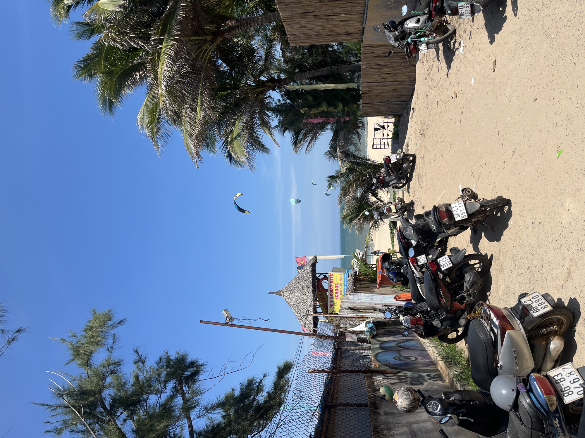 This is an outdoor, eye-level, medium shot of a sandy area near a beach in Phan Thiết, Vietnam. The scene is brightly lit by the sun, with a clear blue sky overhead. Palm trees and other greenery frame the shot.
In the foreground, several motorbikes are parked on the sand. They are facing in various directions, with their wheels resting on the sandy ground. Some of the motorbikes have helmets placed on them.
To the left, a fence made of chain-link and concrete separates the motorbike parking area from a street or pathway. The concrete wall has graffiti on it. A lamppost stands near the fence.
In the mid-ground, a thatched-roof hut with a sign that reads "MUI NE BAY KITE SCHOOL" is visible, indicating a kite surfing school is located here. Further back, the beach is visible with the ocean in the distance. Several kite surfers are seen on the water, with colorful kites in the air.
To the right, a bamboo fence runs along the edge of the sandy area. More motorbikes are parked along this fence.
The overall scene suggests a popular spot for beach activities and water sports in Phan Thiết.