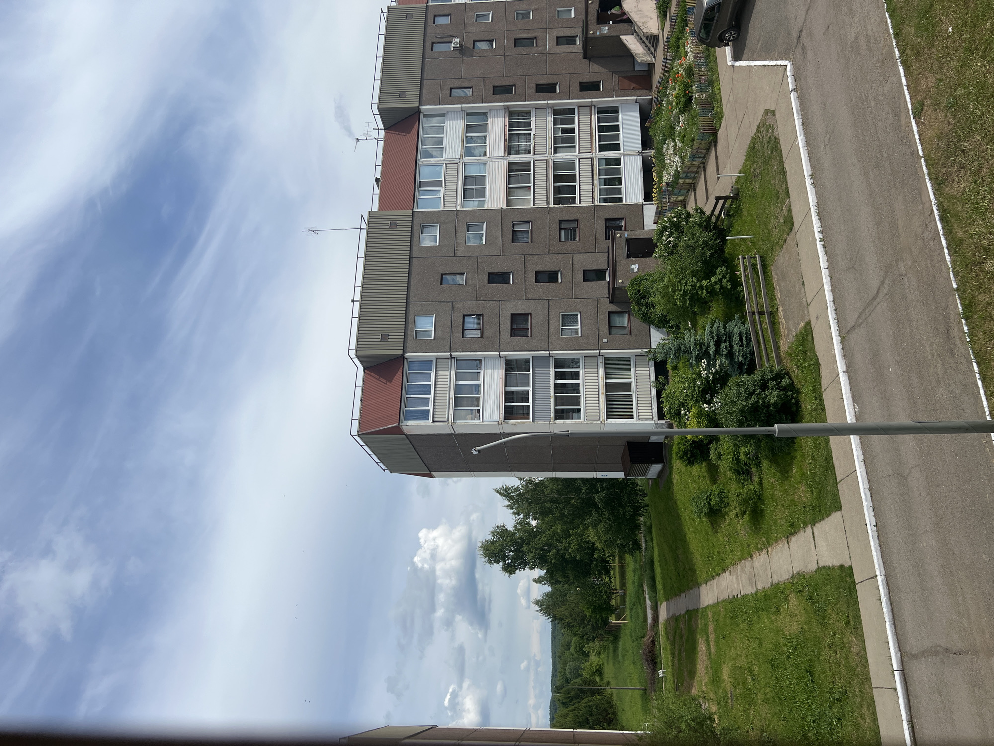 This image captures an outdoor scene dominated by a large, multi-story residential building under a bright, partly cloudy sky. The building, characteristic of Soviet-era architecture, features a grey concrete panel facade with numerous windows and enclosed balconies. Sections of its upper levels and sides are clad in a ribbed, possibly metallic, material, with several antennas visible on the roof.
In the foreground and mid-ground, a green residential area unfolds. This includes well-maintained lawns, various types of bushes, and flower beds bursting with white, yellow, and orange blooms, bordered by a low, colorful wooden fence. Two simple wooden benches are situated among the greenery. A narrow concrete path snakes through the grass, leading towards a wider asphalt road marked by a white line. A dark-colored car is partially visible parked on the asphalt. A tall, grey street lamp post stands upright between the paved path and the road.
Beyond the immediate vicinity of the building and residential landscaping, a natural landscape with dense green trees and distant rolling terrain is visible under the expansive sky. The sky itself is a light blue, streaked with delicate white cirrus clouds and some more defined cumulus formations towards the horizon.
The scene depicts a clear, fair daytime, likely during the warmer months given the lush vegetation. There is no discernible human activity or visible text. The setting is consistent with a residential block in Марковский, Russia.