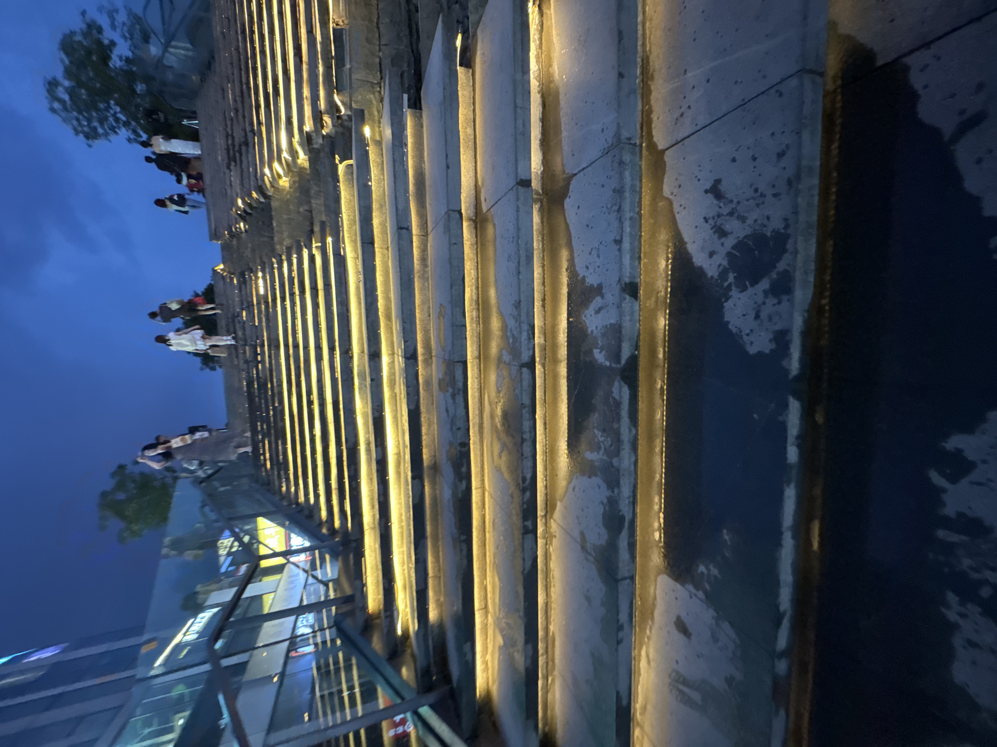 The image shows a wide, stone staircase illuminated by yellow lights embedded along the risers, leading upwards. The sky is dark blue, indicating it is dusk or night. Several individuals are visible walking up the stairs or standing at the top. On the left side of the stairs, a modern building with glass windows reflects the lights. The stone steps appear wet, with puddles of water and reflections. A metal railing runs along the left side of the staircase. This scene is located at the entrance to the Olympic Park in Chaoyang District, China, a public space known for its impressive architecture and open areas.