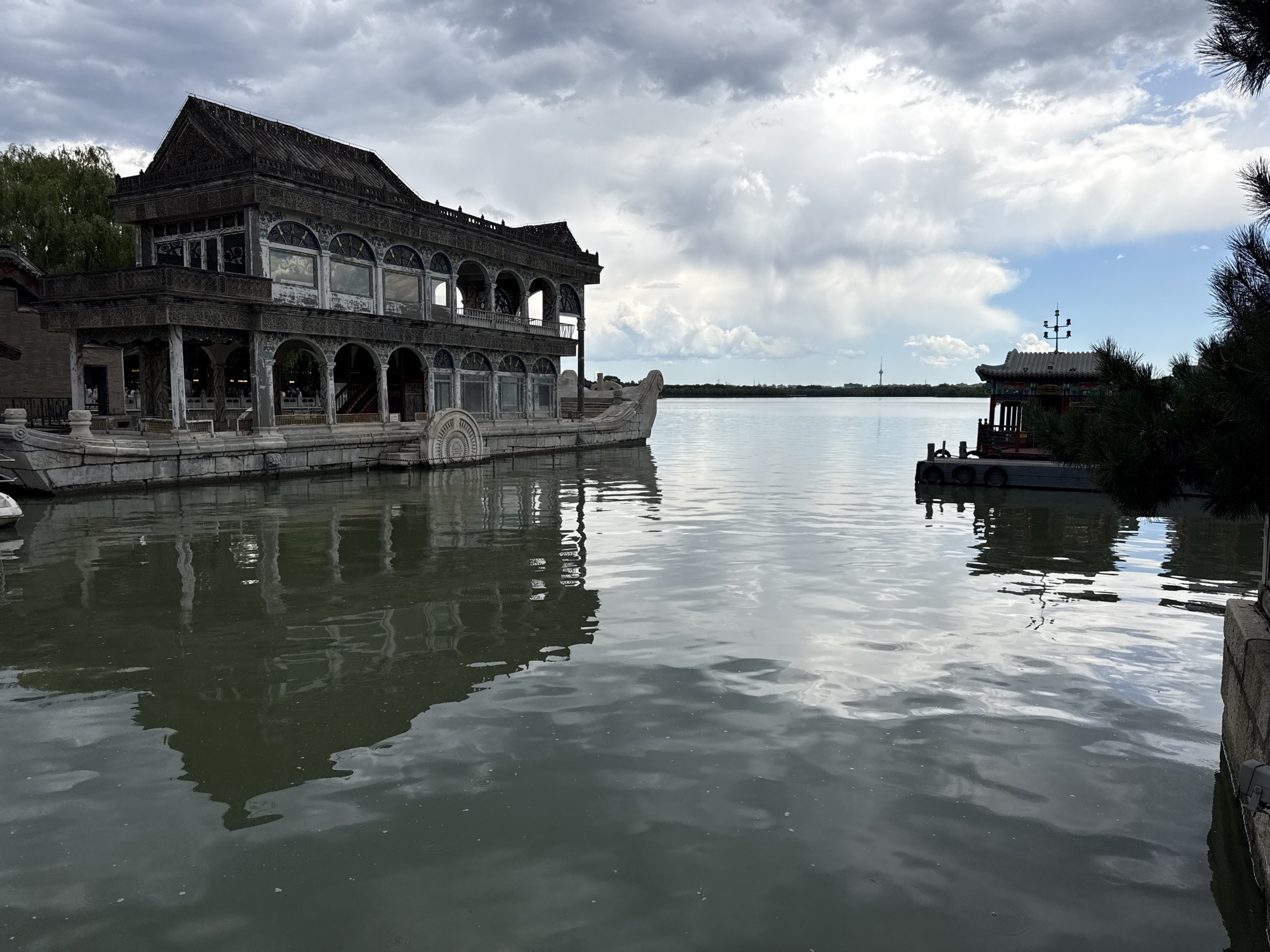The image depicts the Marble Boat (Qingyanfang) on Kunming Lake within the Summer Palace in Haidian District, Beijing, China. A two-story stone structure designed to resemble a boat occupies the left and central midground. This structure features traditional Chinese architectural elements, including a tiled roof, ornate carvings, arched windows on the lower level, and rectangular windows on the upper level. Its right end terminates in a decorative scroll-like prow.
The water of Kunming Lake fills the foreground and midground, exhibiting a smooth, reflective surface with subtle ripples, mirroring the sky and architectural elements. To the far right, a smaller, traditional Chinese pavilion-like structure with a curved, multi-colored roof and a decorative lamppost on its peak is situated on a dock with circular black fenders. This pavilion is partially framed by dark green conifer foliage in the extreme right foreground.
The sky above is predominantly overcast with white and grey clouds, though distinct patches of blue sky are visible, particularly towards the distant horizon. A low treeline is present along the distant shoreline, and a slender communication tower (resembling the Central Radio and TV Tower) is discernible against the sky in the far distance on the right side. Green deciduous foliage, likely trees, is visible behind the Marble Boat on the left. Stone embankments line the immediate right edge of the water. No individuals are present in the scene.