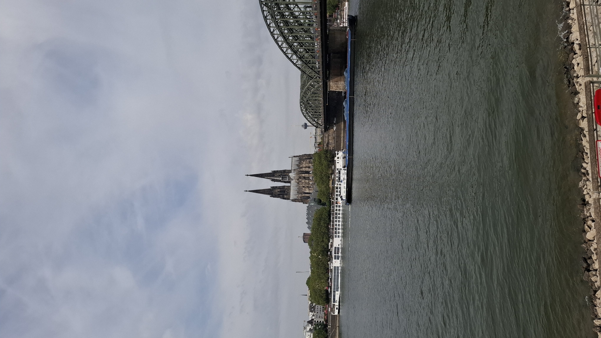 The image depicts a wide river with two barges, one white passenger vessel and one dark cargo vessel, traversing its waters. In the background, the iconic Cologne Cathedral with its twin spires dominates the skyline, accompanied by a large, green arched bridge spanning the river. The cityscape on the left features buildings and trees, while the right side shows the bridge and a tower. The sky above is overcast with clouds. The foreground features a rocky embankment lining the river. The scene is a view of the Rhine River in Cologne, Germany, with the Hohenzollern Bridge and Cologne Cathedral prominently featured.