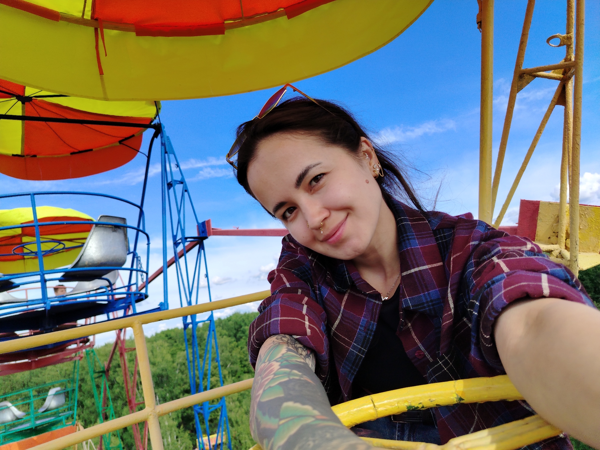 A young woman is captured in a selfie taken on an amusement ride, likely a Ferris wheel, in Chaikovsky, Russia. She is positioned in the lower right foreground, looking towards the camera with a slight smile. Her dark hair is pulled back, with sunglasses perched on top of her head. She wears multiple ear piercings, including hoops and studs, along with two nose rings and a nose stud. She is dressed in a dark top underneath a long-sleeved plaid shirt featuring shades of red, blue, and white. A prominent, colorful floral or leafy tattoo is visible on her left forearm, extending from her wrist.
The background reveals the colorful, aged metal framework of the amusement ride, painted in bright yellow, orange, red, and blue. Elements such as railings and parts of passenger capsules are visible, suggesting motion or height. The sky above is a clear, vibrant blue with wispy white clouds, indicating a bright and sunny daytime setting. In the distance, a dense expanse of green trees and foliage suggests a park or natural landscape surrounding the amusement area. The perspective of the image suggests it was taken from inside one of the ride's compartments, looking out. No visible text is present in the image.
