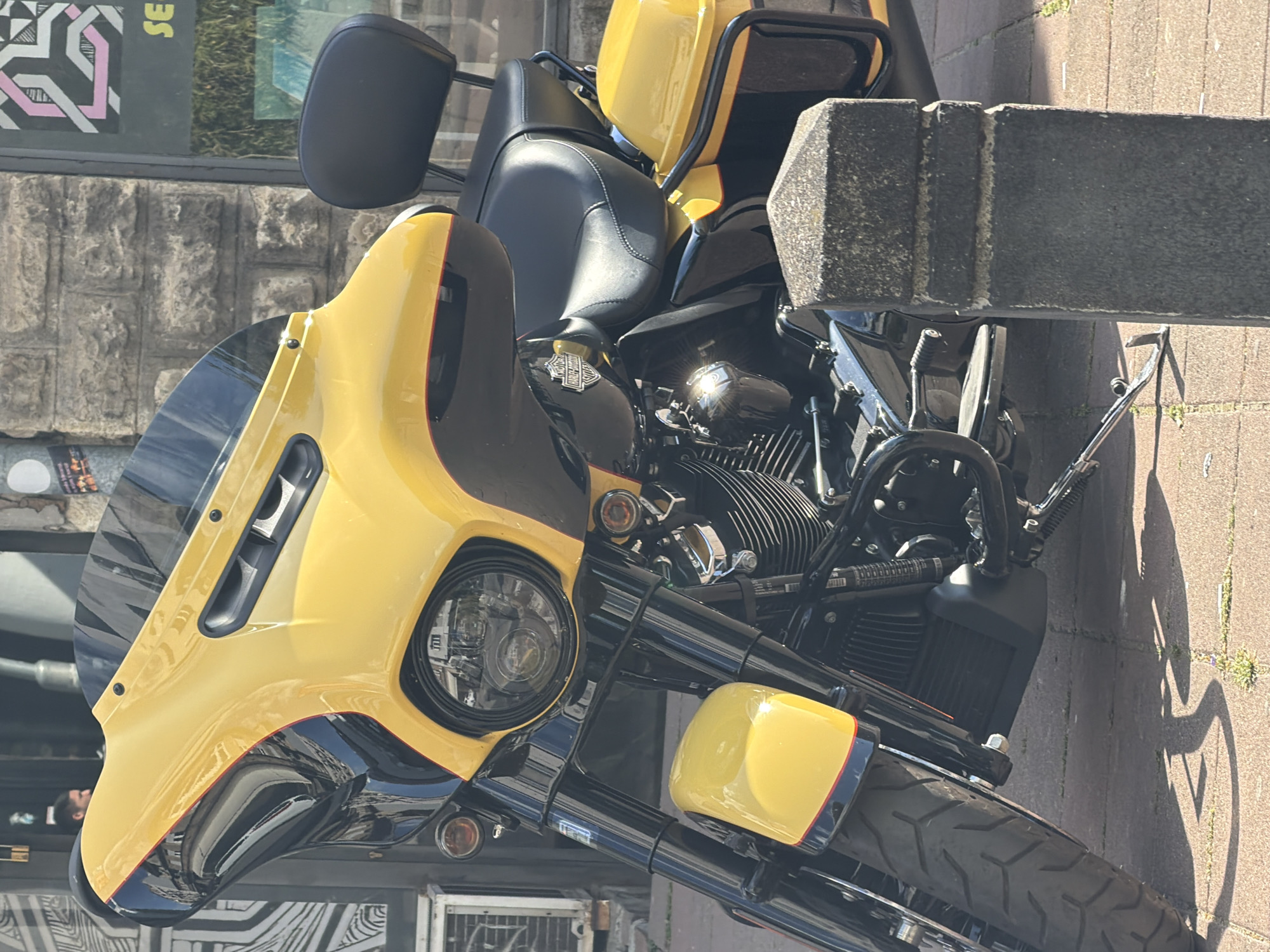 The image shows a yellow and black Harley-Davidson motorcycle parked on a brick sidewalk, close to a gray concrete street fixture. The motorcycle is oriented diagonally in the frame. The front of the bike features a large yellow fairing with a windshield and a round headlight. The engine and frame are predominantly black with chrome accents. The back of the bike has a black seat, a black backrest, and yellow saddlebags. The background suggests a city environment, with a building visible, including graffiti on the building. Belgrade, Serbia is not identifiable.