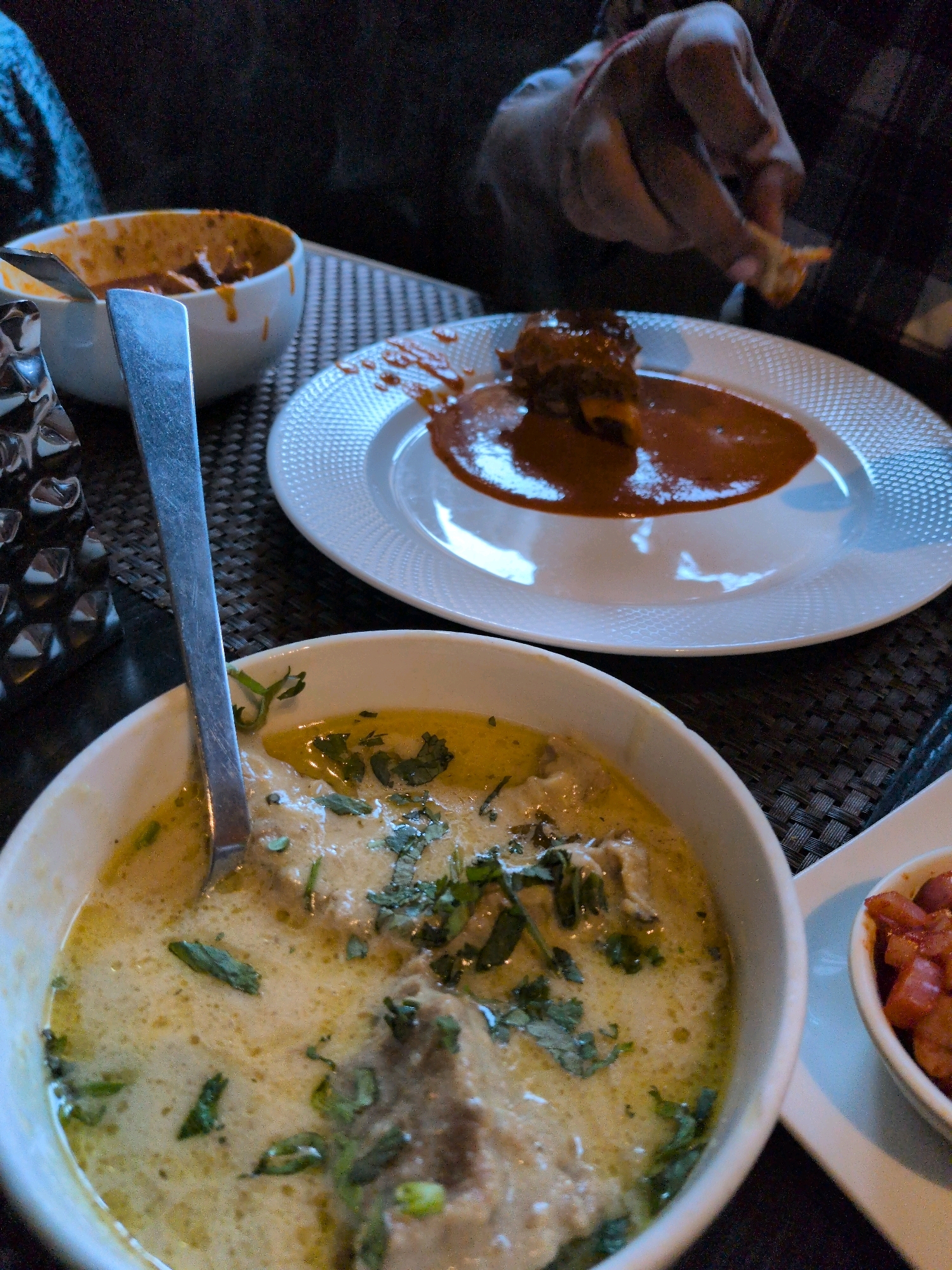 In Srinagar, India, a close-up shot captures a dining scene. In the foreground, a white bowl filled with a creamy, light-colored curry, likely a Kashmiri delicacy like Rogan Josh or Chicken Korma, is visible. The curry is garnished with chopped green herbs, possibly cilantro. A silver spoon rests within the bowl. To the right, a smaller white bowl contains a red, diced food item, possibly tomatoes or other vegetables. In the background, a white plate holds a piece of meat in a thick, reddish-brown sauce. A person's hand, with fingers stained a light orange, is seen reaching towards the plate, about to pick up a piece of food with their fingers. The background is dark, suggesting an indoor setting with low lighting, typical of a restaurant ambiance. The table is covered with a textured mat. The overall impression is a depiction of a meal being enjoyed, showcasing a portion of Indian cuisine.