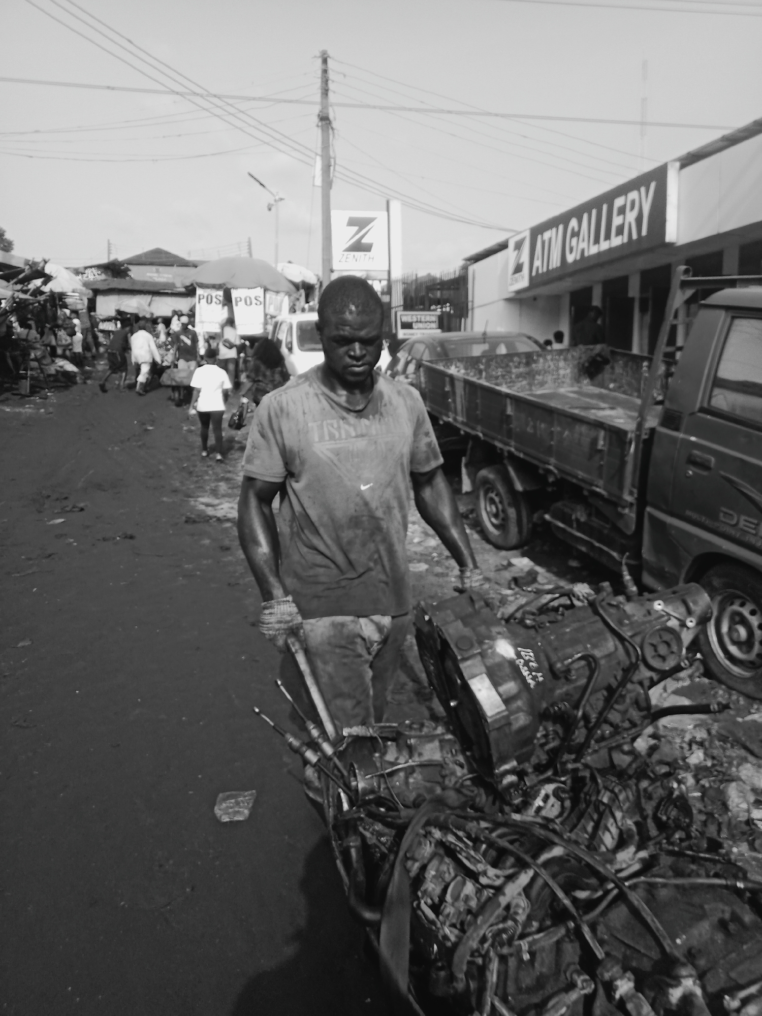 A black and white photograph depicts a man standing in what appears to be a marketplace in Onitsha, Nigeria. He is hauling a large, disassembled machine, possibly an engine, using a hand truck. The man is wearing a t-shirt and appears to be working in a dirty or greasy environment. Behind him, there is a bustling marketplace with people walking around, vendors with stalls, and vehicles. In the background, signs for "ZENITH" and "ATM GALLERY" are visible, as well as power lines overhead. The road is uneven and appears muddy.