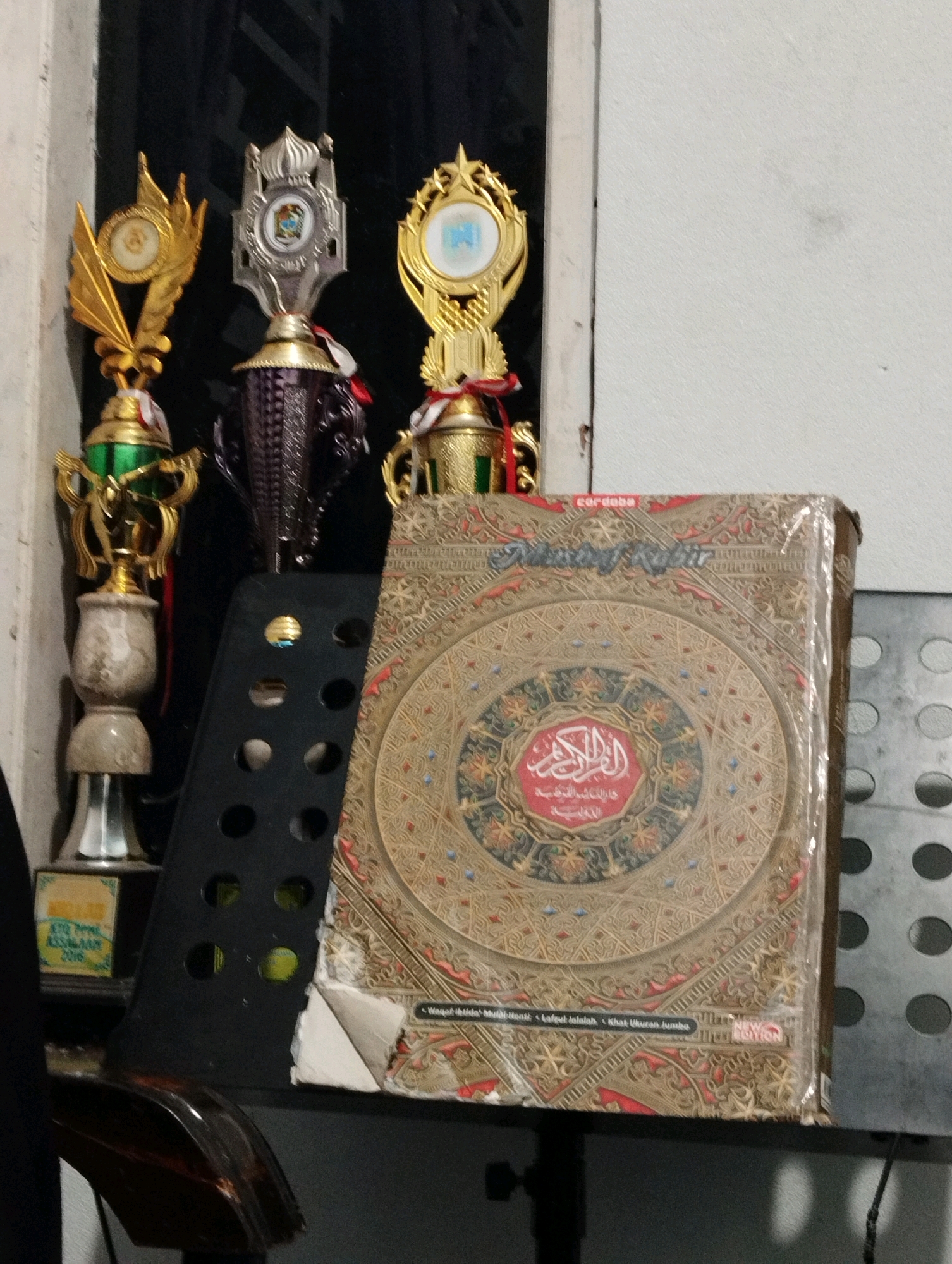 This is a medium shot, interior, eye-level, medium-angle photograph featuring a collection of trophies and a book. The background appears to be a window and a wall. The lighting is dim, suggesting it was taken indoors at night or in a poorly lit room.
In the foreground, to the left, are three trophies. The leftmost trophy is gold with green and marble accents. The middle trophy is silver and purple. The rightmost trophy is gold with red and white ribbon accents. To the right of the trophies, a book with intricate gold and red patterns is displayed. The book appears to be a Quran, with Arabic calligraphy visible on its cover. The book is wrapped in clear plastic. A black music stand is partially visible behind the trophies and book, with several circular holes.
In the lower left corner, a small yellow and blue award is visible. The inscription on this award appears to read "ASTRA PEAL ASSESMENT 2018". The overall scene suggests a place where awards have been received and religious texts are kept, potentially a personal study or a communal space in a religious institution. The presence of the music stand might indicate a connection to musical activities or performances.
The image does not provide specific visual cues to pinpoint the location within Salatiga, Indonesia.
