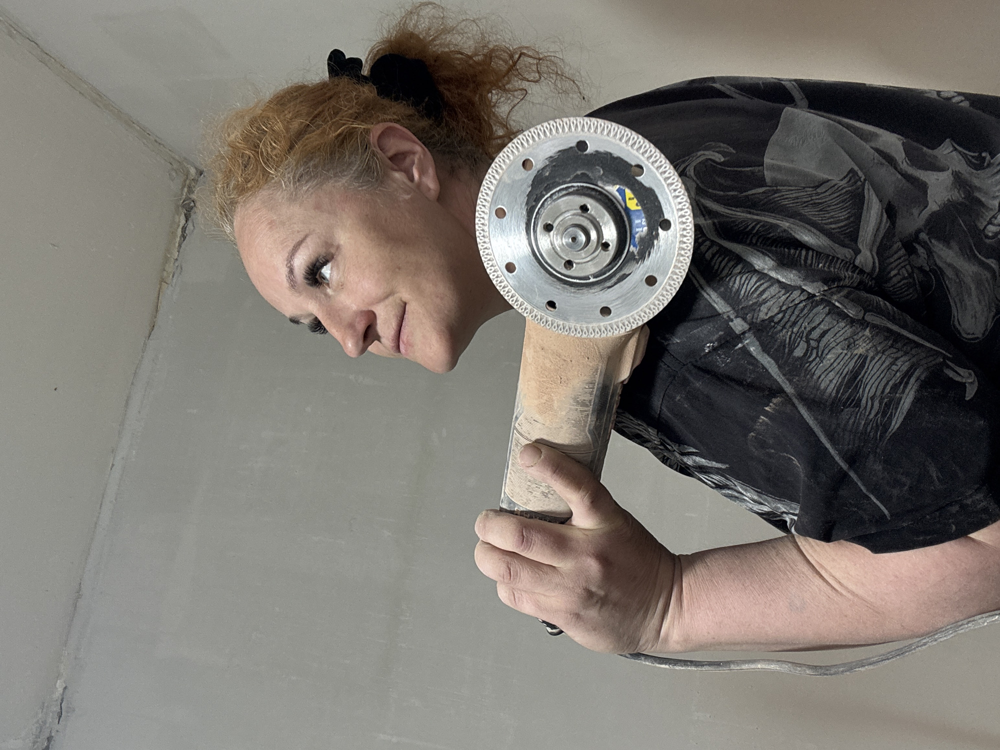 A woman is holding an angle grinder in what appears to be an indoor location, possibly a room in a house. The woman is wearing a black t-shirt, her hair is red, and she has it pulled back in a ponytail with a black scrunchie. The angle grinder has a metal blade. The background is a light-colored wall, and a corner is visible. The location of the image is unknown, therefore, it cannot be verified that the image was taken in Chaikovsky, Russia.
