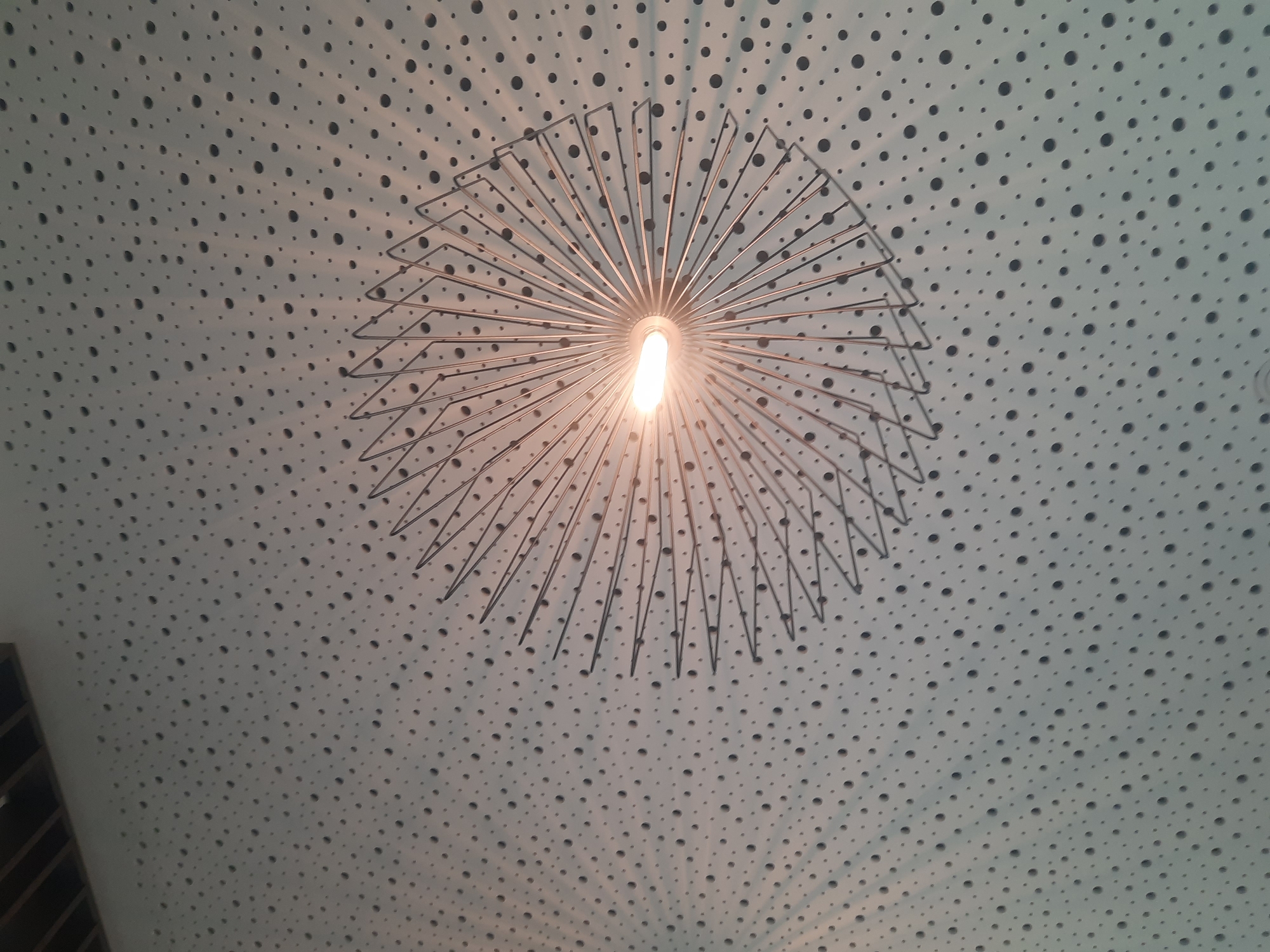The image shows a ceiling with a light fixture. The ceiling is white and has a pattern of small black dots. The light fixture is in the center of the image and is a starburst shape made of metal rods with a fluorescent bulb in the middle. The light is on and is casting shadows on the ceiling. The image appears to have been taken indoors, likely in a building.