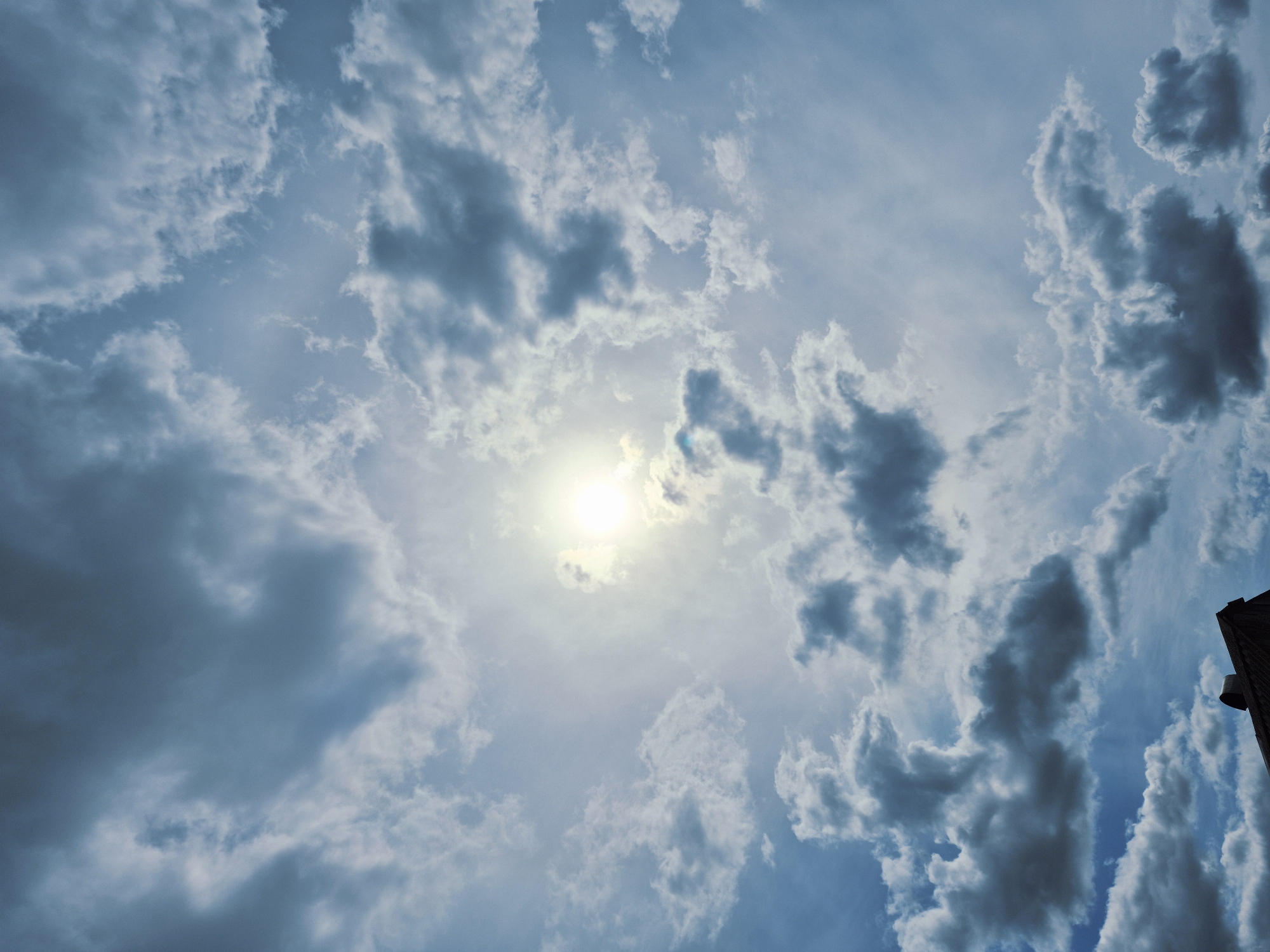 This is a daytime upward-facing shot of a sky filled with various cloud formations. The sun, a bright luminous disc, is centrally positioned towards the upper part of the frame, partially obscured by a thin veil of clouds.
The sky displays a range of blue tones, from a lighter, almost ethereal blue around the sun to deeper, more saturated blues where the sky is clearer. The clouds are diverse in shape and density; some are wispy and translucent, allowing sunlight to pass through, while others are thicker, darker, and more defined, casting subtle shadows and adding depth to the scene. These more robust clouds are particularly prominent on the right side of the image, appearing almost sculptural against the lighter background.
A small portion of a dark, vertical structure, possibly part of a building or a tall pole, is visible along the extreme right edge of the frame, suggesting an urban or constructed environment in the general vicinity. No people or specific activities are depicted, and no visible text is present. The image conveys a sense of open space and the dynamic beauty of natural light and atmospheric conditions in Дойная, Russia.