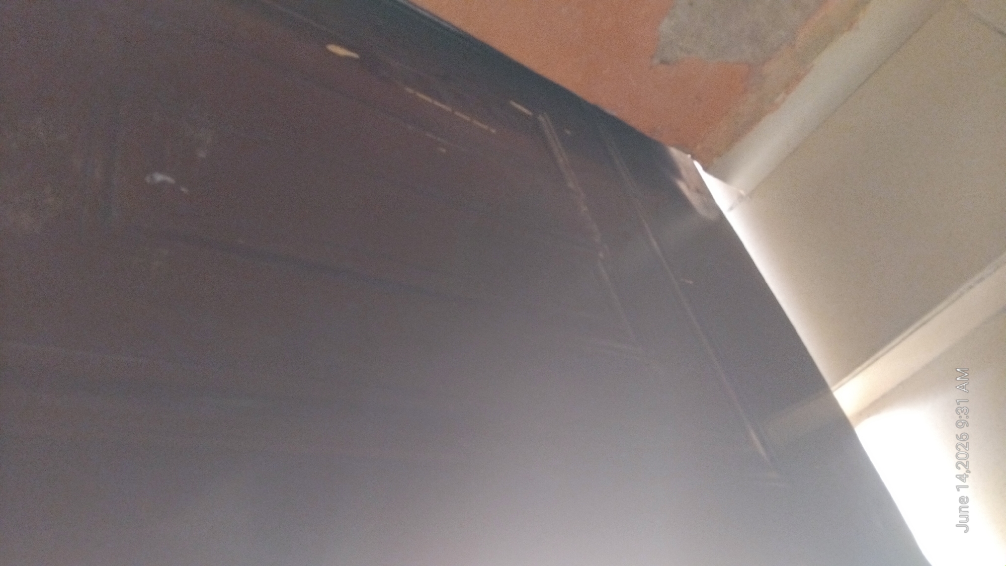 The image shows a close-up view of the top of a dark-colored door and a portion of an orange-colored wall, likely inside a building. The door appears to have some faint lines or grooves on its surface. There are a few small white specks on the door, possibly paint or dust. The orange wall is rough in texture and shows some lighter colored patches. The bottom of the image features a bright, possibly sunlit, area with a light-colored surface. A timestamp in the bottom left corner reads "June 14, 2026 9:31 AM". The image was taken from a low angle, looking upwards. There are no people or discernible actions depicted in the image. The scene suggests an indoor environment, possibly a room or hallway, in Jalingo, Nigeria, based on the provided location.