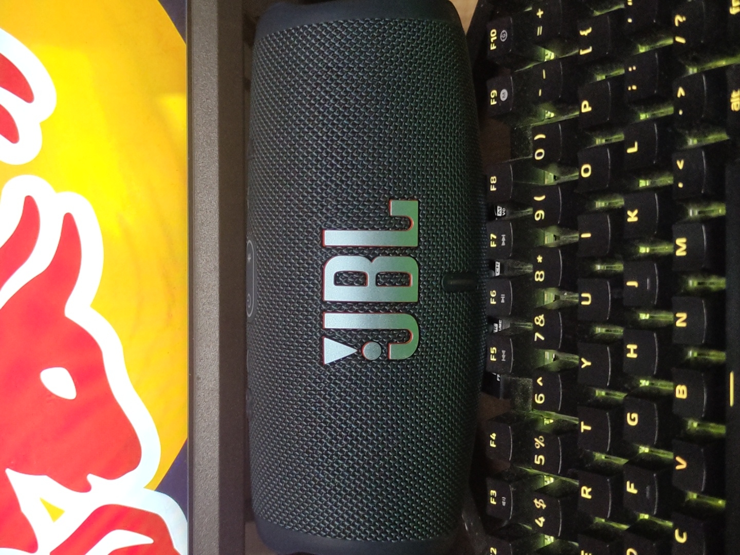 An overhead, vertically oriented shot captures a JBL portable speaker positioned between a mechanical keyboard and a partial Red Bull logo, likely on a desk or table. The scene appears dimly lit, suggested by the bright yellow-green backlighting of the keyboard keys. The items are arranged in a horizontal line across the frame, with the speaker central.
The JBL speaker is black and cylindrical, featuring a textured mesh exterior. Its prominent "JBL" logo, in a light green or teal color, is displayed prominently in the center. To the right of the speaker is a black mechanical keyboard with individually backlit keys. The backlighting casts a yellowish-green glow, illuminating key legends such as F5 through F10, numbers 0-9 with corresponding symbols like '&', '*', '(', ')', and letters including P, L, K, J, U, Y, T, R, G, F, B, N, M, C. Modifier keys like "alt" are also visible. To the left of the speaker, a bright yellow surface with a partial red and white logo is visible. This is clearly identifiable as part of a Red Bull energy drink logo, showing the head and partial body of one of the iconic red bulls.
The image depicts a personal computing and entertainment setup. While no specific activity is shown, the arrangement suggests a space for listening to music while working or gaming. The dim lighting and keyboard backlighting hint at evening or nighttime use, or an indoor setting with low ambient light. The location is reportedly General Toshevo, Bulgaria.