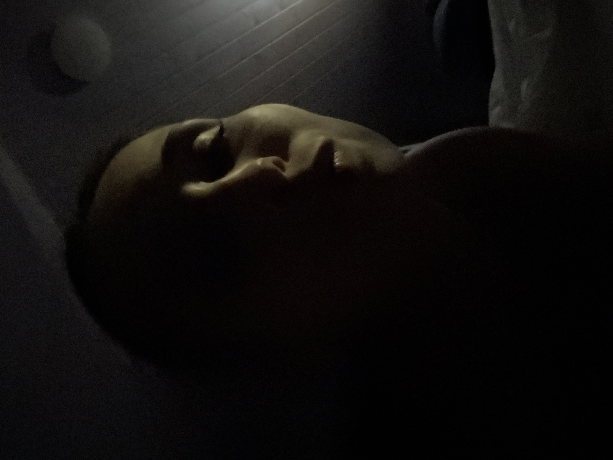 A low-angle, close-up, dimly lit indoor shot features a person's face from the side. The light source appears to be above and to the right, casting a strong shadow over the left side of the face. The person's right eye is visible, and their lips are slightly parted. The background consists of a ribbed ceiling and a circular light fixture. A white surface, possibly a bed or pillow, is visible in the lower right corner. The image is dark and somewhat blurry, making it difficult to discern fine details.