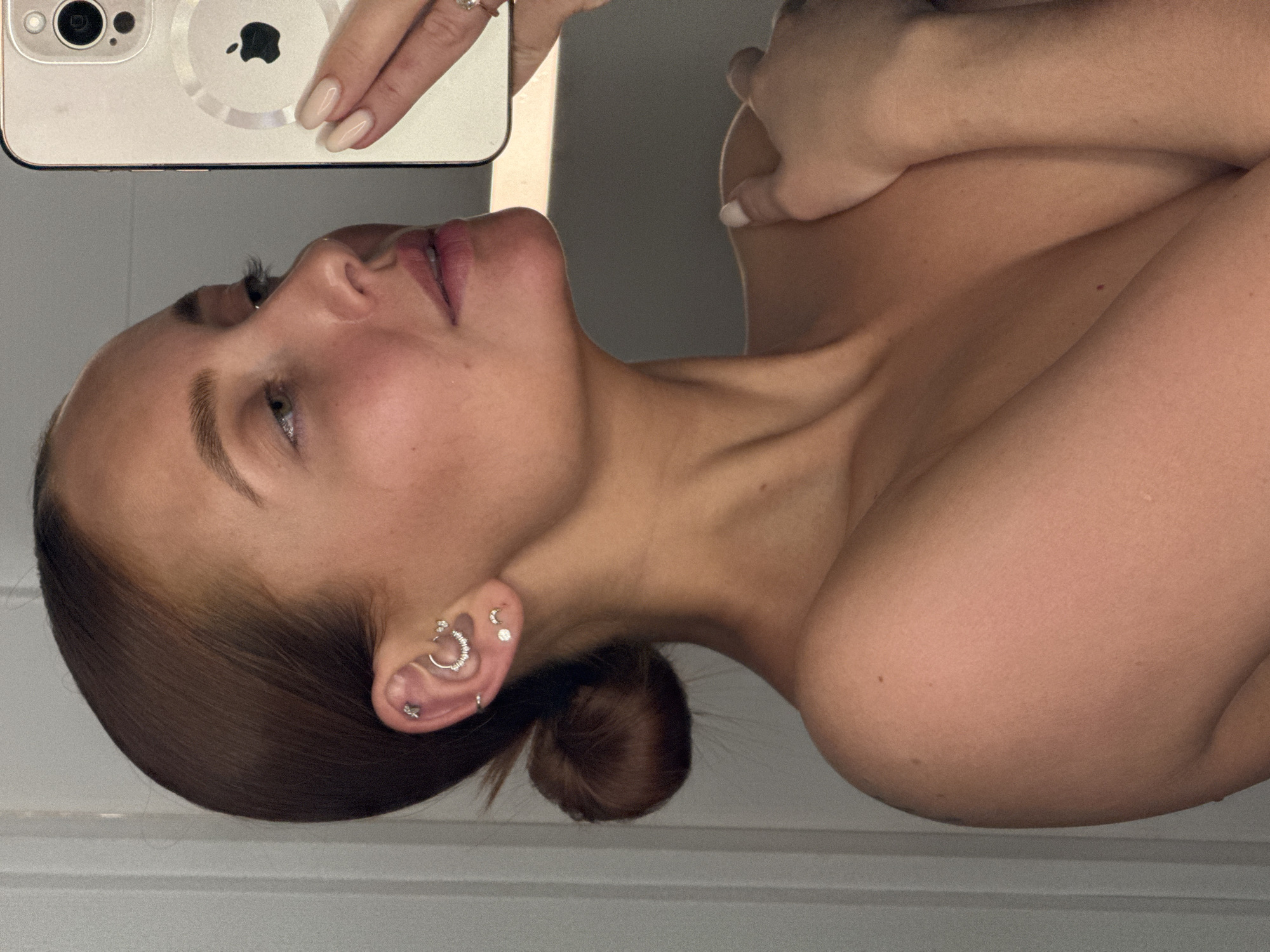 A woman is taking a selfie. She is holding a white iPhone in her left hand, which is positioned near the top left of the frame. Her head is tilted back, her face angled towards the camera. She has light brown hair pulled back into a bun, visible on the left side of her neck. She has multiple piercings in her left ear. She is facing away from an unidentified, light-colored surface. The setting and specific location within the city of Chaikovsky, Russia, are not discernible from the image.