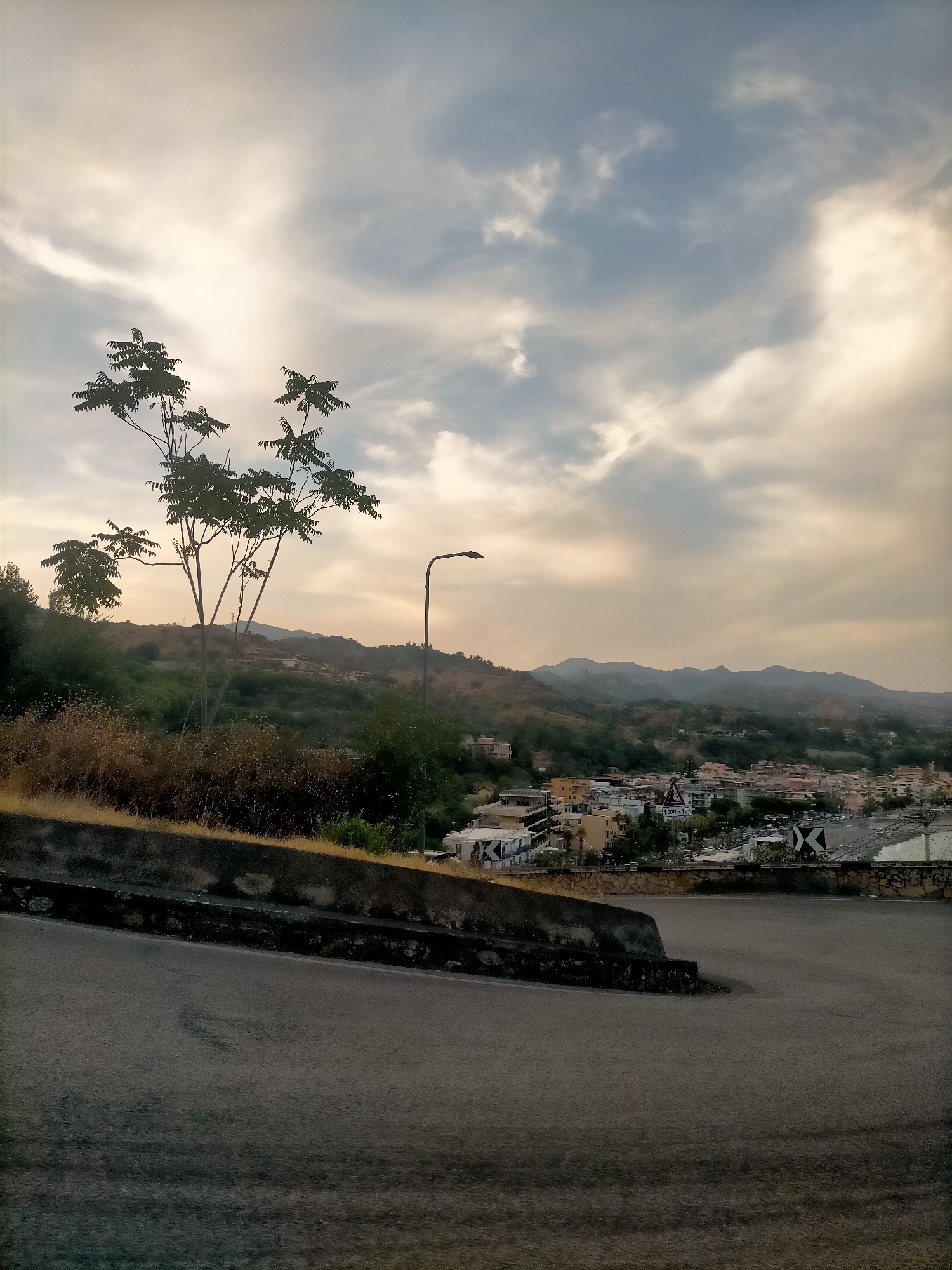 The image shows an elevated view of a coastal town nestled against rolling hills under a cloudy sky. In the foreground, a dark asphalt road curves to the right, with a concrete barrier and dry vegetation along the left side. Beyond the barrier, a small tree with sparse foliage stands out against the background. Further in the distance, the town's buildings, a mix of light-colored structures, are visible, some with balconies and tiled roofs. A hint of a beach or waterfront area can be seen to the right. The overall lighting suggests it might be late afternoon or early morning, with a soft, diffused light. Mountains form the backdrop, disappearing into a hazy horizon. A street lamp is visible on the left side of the frame, and traffic signs are present in the middle distance.
