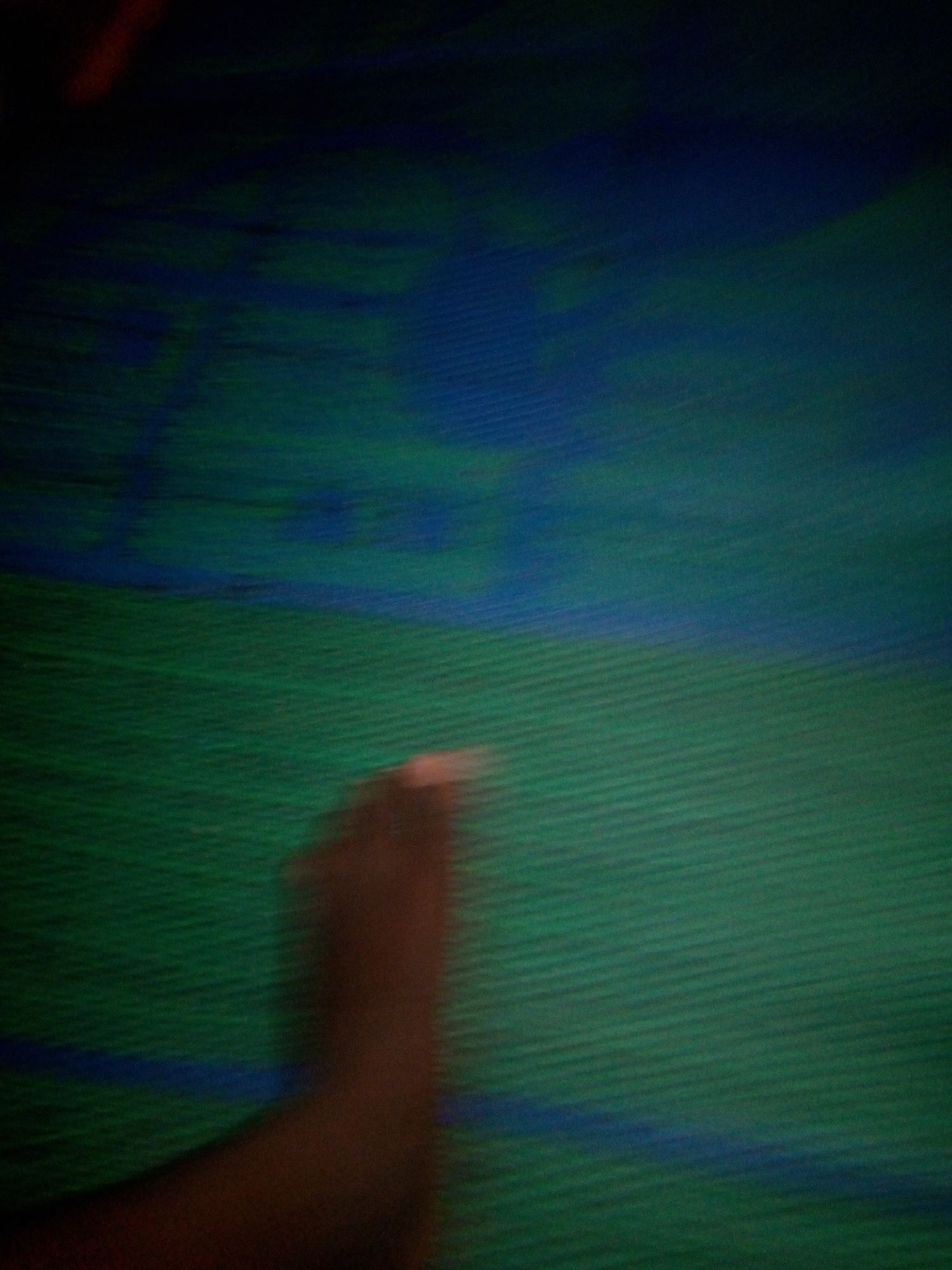 The image shows a blurry, low-light scene with a predominantly green and blue color palette. In the foreground, a dark brown, blurred shape, possibly a hand or arm, is visible. The background appears to be a textured surface, possibly fabric or a mat, with horizontal lines. There are also streaks of blue within the green texture. The overall impression is one of movement or a shaky camera. The context of Jalingo, Nigeria, is not evident from the visual information presented in the image.