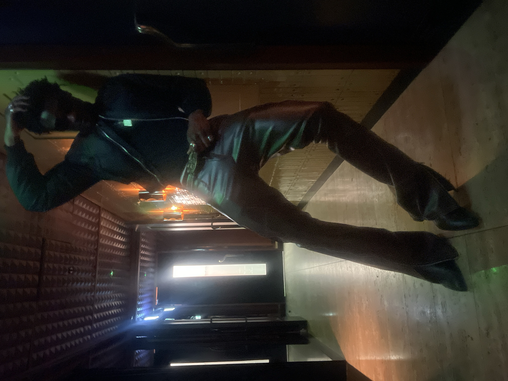 A person with dark hair is standing in a hallway, wearing a black shirt and brown pants. The hallway has textured walls on the left and smooth walls on the right, with a bright light emanating from the end of the hall. The person is holding their head with their left hand, and their right hand is near their waist. The floor appears to be made of light-colored tiles.