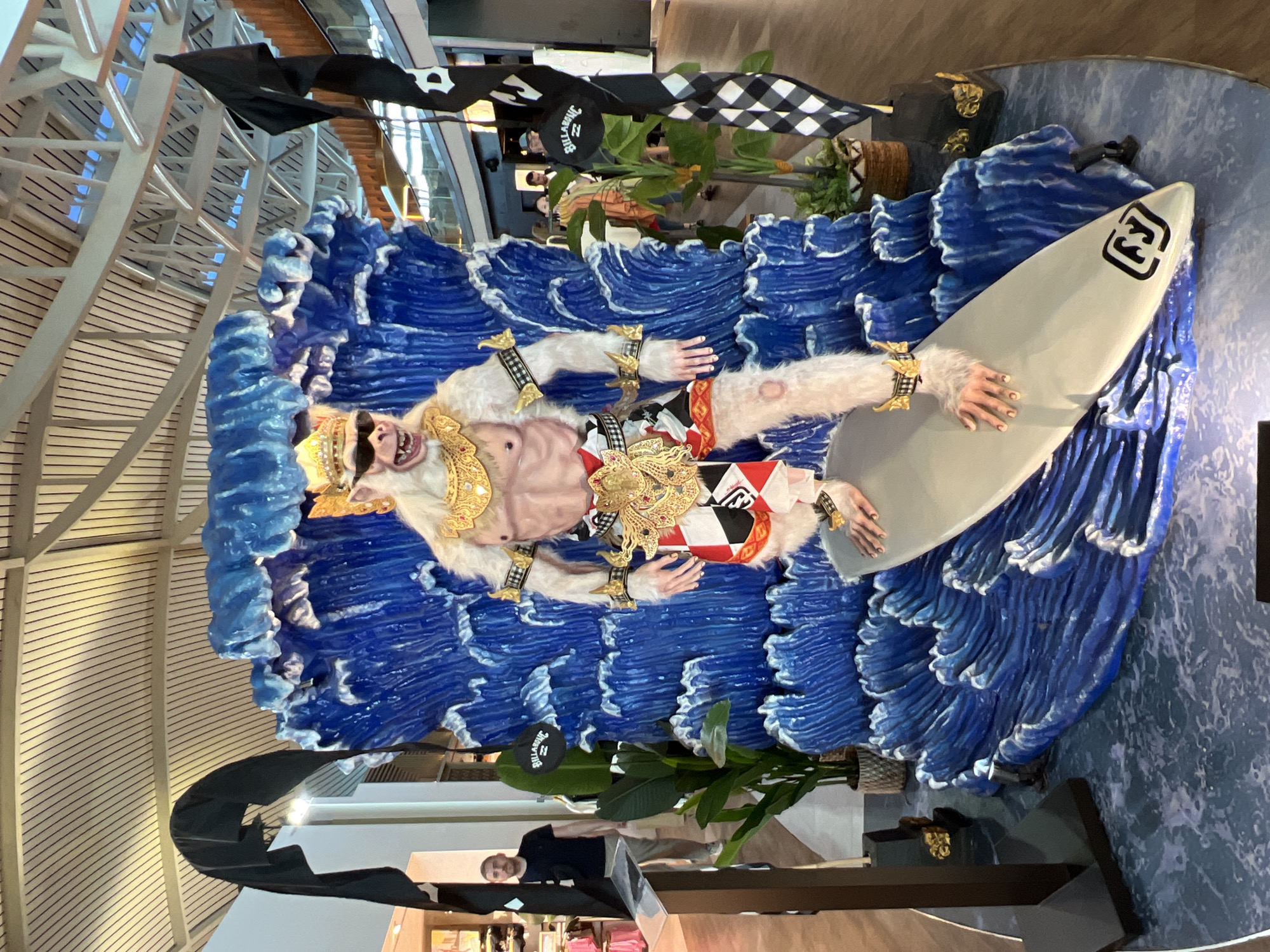 The image depicts a large, theatrical display featuring a life-sized statue of a white monkey character dressed as a surfer. The statue is positioned on a surfboard that appears to be riding a stylized, bright blue wave. The monkey is wearing a golden crown, sunglasses, ornate gold jewelry on its neck, wrists, and ankles, and a red, white, and black patterned sarong. The background of the display is a large, textured blue wave.
Flanking the display are tall, black flags with white checkered patterns and a black flag with a white logo. To the left of the display, a man wearing a dark t-shirt and light-colored pants is visible, standing and looking towards the display. Behind the man, there are shops with clothing racks. To the right, there are potted plants and more flags. In the background, there are escalators, railings, and people moving about, suggesting an indoor public space like a mall or an airport terminal.
The scene is likely set up as a promotional or decorative installation, possibly related to surfing culture or a local event. Given the location context provided as Kuta, Indonesia, this display could be found in a shopping center or a tourist-oriented area within the city, which is well-known for its beaches and surfing. The lighting suggests an indoor environment with artificial light sources overhead. The overall presentation is colorful and eye-catching, designed to attract attention.