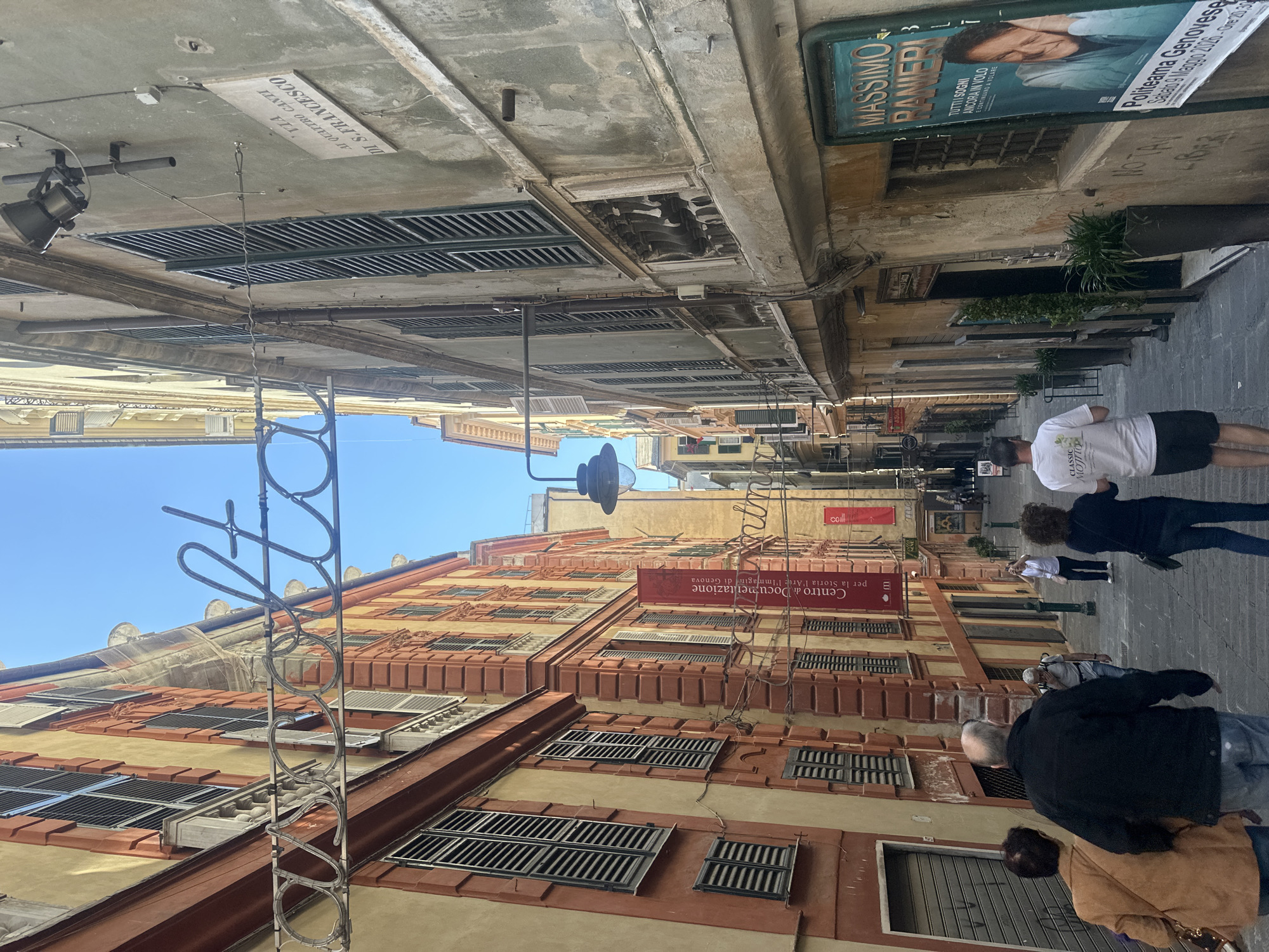 The image depicts a bustling, narrow street or alleyway in Genoa, Italy, illuminated by bright daylight under a clear blue sky. The scene is viewed looking down the street, with tall, multi-story buildings lining both sides, characterized by their aged, textured facades in warm tones of yellow, orange, and terracotta, featuring numerous windows, many fitted with closed dark green or brown louvered shutters.
Several individuals are present, mostly seen from behind, walking away from the camera or standing. In the foreground, a person in a black jacket and blue jeans, accompanied by another in a brown jacket, walks right. Further down the street, a person in a white t-shirt and black shorts, with the text "CLASSIC MOJITO" visible on their back, also walks away. Other people are scattered along the street, suggesting everyday pedestrian activity or tourism.
Overhead, a complex network of wires and light fixtures spans the gap between the buildings. A prominent, ornate metal structure, possibly part of a decorative sign or lighting, extends into the sky on the left side of the foreground.
Notable textual details include several signs and posters:
* A street sign mounted high on a building reads, "VIA AI QUATTRO CANTI DI S. FRANCESCO".
* A large poster advertises "MASSIMO RANIERI" with the show title "TUTTI SOGNI ANCORA IN VOLO E CONTINUANDO A VOLARE", specifying "Politeama Genovese Sabato 9 Maggio 2020 - Ore 20:30".
* A red banner on a building facade states, "Centro di Documentazione per la Storia l'Arte l'Immagine di Genova".
* Graffiti that appears to read "NOTAY" is visible on a wall.
The time of day is clearly sunny and daytime, with no shadows suggesting late afternoon or early morning. The architecture and street layout are typical of historic European city centers, consistent with the Genoa, Italy context.