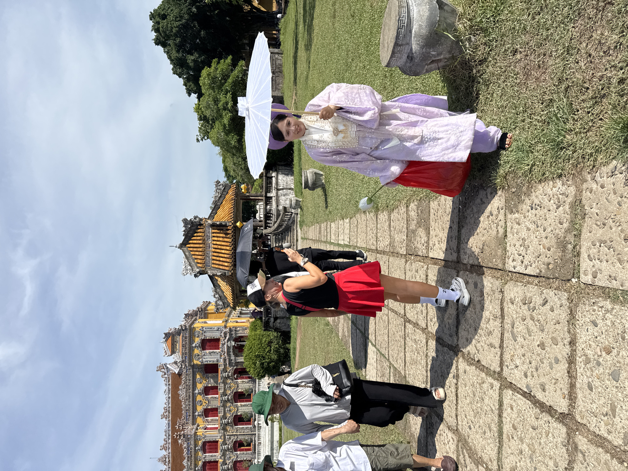 In Phú Xuân Ward, Vietnam, a group of tourists walks along a stone pathway in a grassy courtyard. A woman in a traditional light purple robe, holding a white parasol and a red bag, walks towards the camera. To her left, a woman in a red skirt and black top, wearing a baseball cap, is taking a photo with her phone. Further left, two individuals, one wearing a green hat and the other in a grey jacket, walk hand-in-hand. In the background, a large, ornate yellow building with red windows is visible, along with a traditional tiled roof structure and lush green trees under a bright, cloudy sky. Several large, stone urns are placed on the grass.