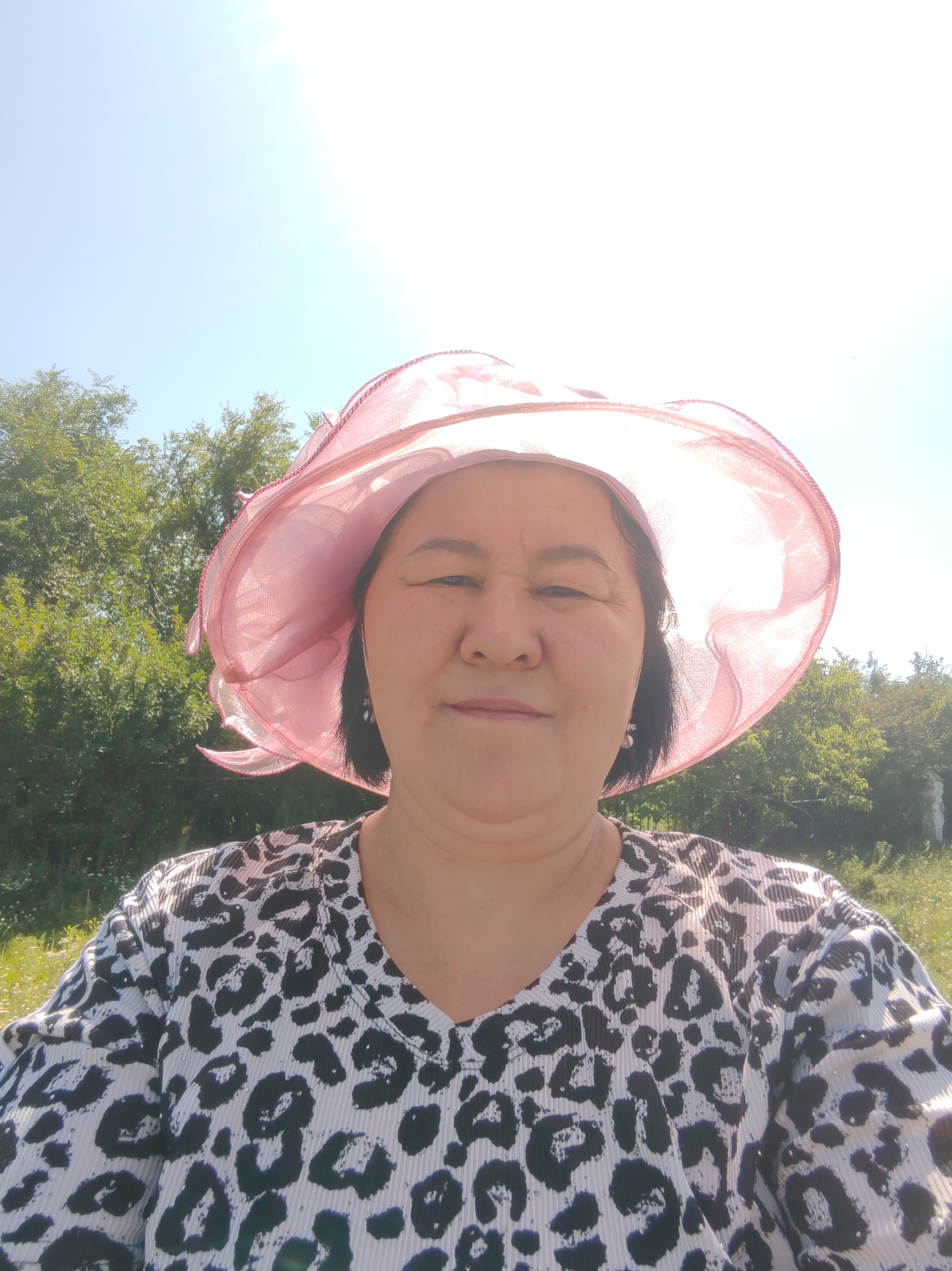 A woman is captured in a close-up, front-facing selfie. She is wearing a large, sheer pink hat that casts a shadow on her face. Her patterned shirt has a leopard-like print in black and white. The background is outdoors, with lush green trees and foliage under a bright, sunny sky, suggesting a natural, possibly rural setting. The image appears to be taken in or near the city of Akzhar, Kazakhstan, given the provided context.