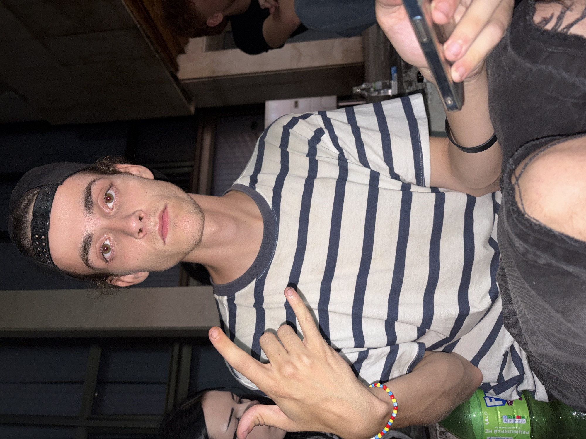 The image depicts a group of young people outdoors at night. The primary subject is a young man in the foreground, wearing a black baseball cap, a striped t-shirt, and dark, ripped pants. He is making a "rock on" hand gesture. To his left, a young woman is partially visible. In the background, another person with reddish hair is seated. A green plastic bottle of what appears to be a beverage is on the ground to the left of the primary subject. A smartphone is held by a person whose hand is visible in the lower right of the frame. The background suggests an urban setting with buildings and low lighting. The presence of concrete structures and a somewhat derelict appearance of the surroundings might indicate an industrial or less maintained area within the city of Vratsa, Bulgaria.