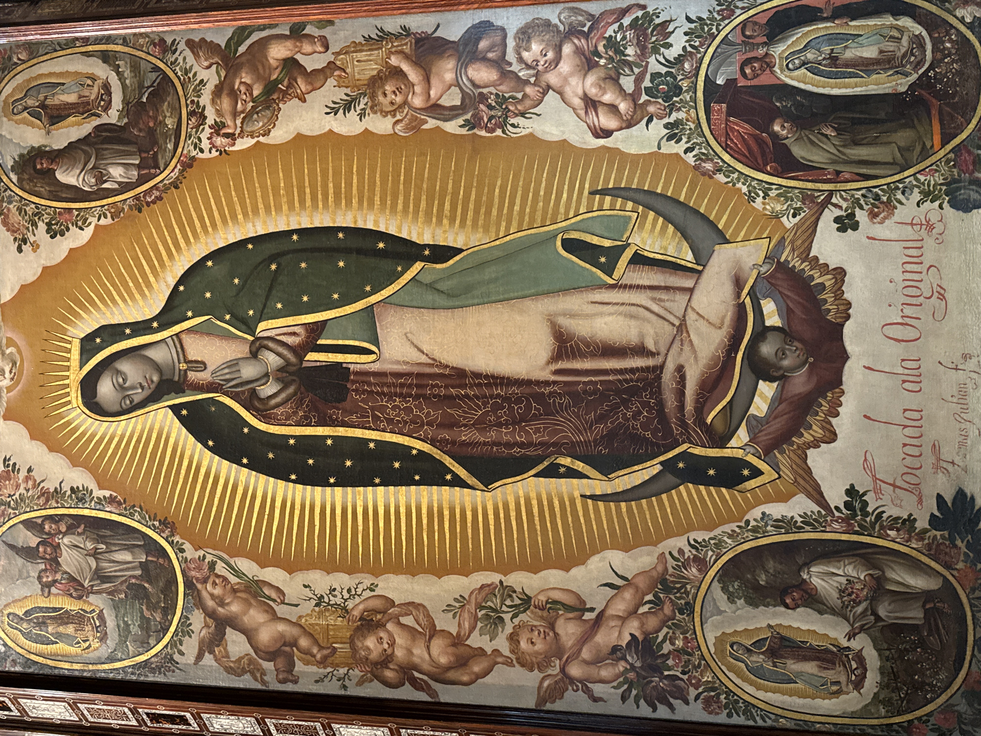 This is a detailed artistic rendition of the Virgin of Guadalupe, likely a painting. The Virgin is depicted with her characteristic dark green cloak adorned with gold stars, set against a backdrop of golden rays emanating from her. Her hands are clasped in prayer, and she stands upon a crescent moon, supported by an angel. The artwork is further embellished with cherubic figures and framed vignettes illustrating scenes related to the Virgin of Guadalupe's story. In the lower right corner, there is text that reads "Locada ala Original Fomas Zulian, fir." The image appears to be a historical or religious painting, possibly located in a church or museum in Mexico City, Mexico, considering the subject matter.