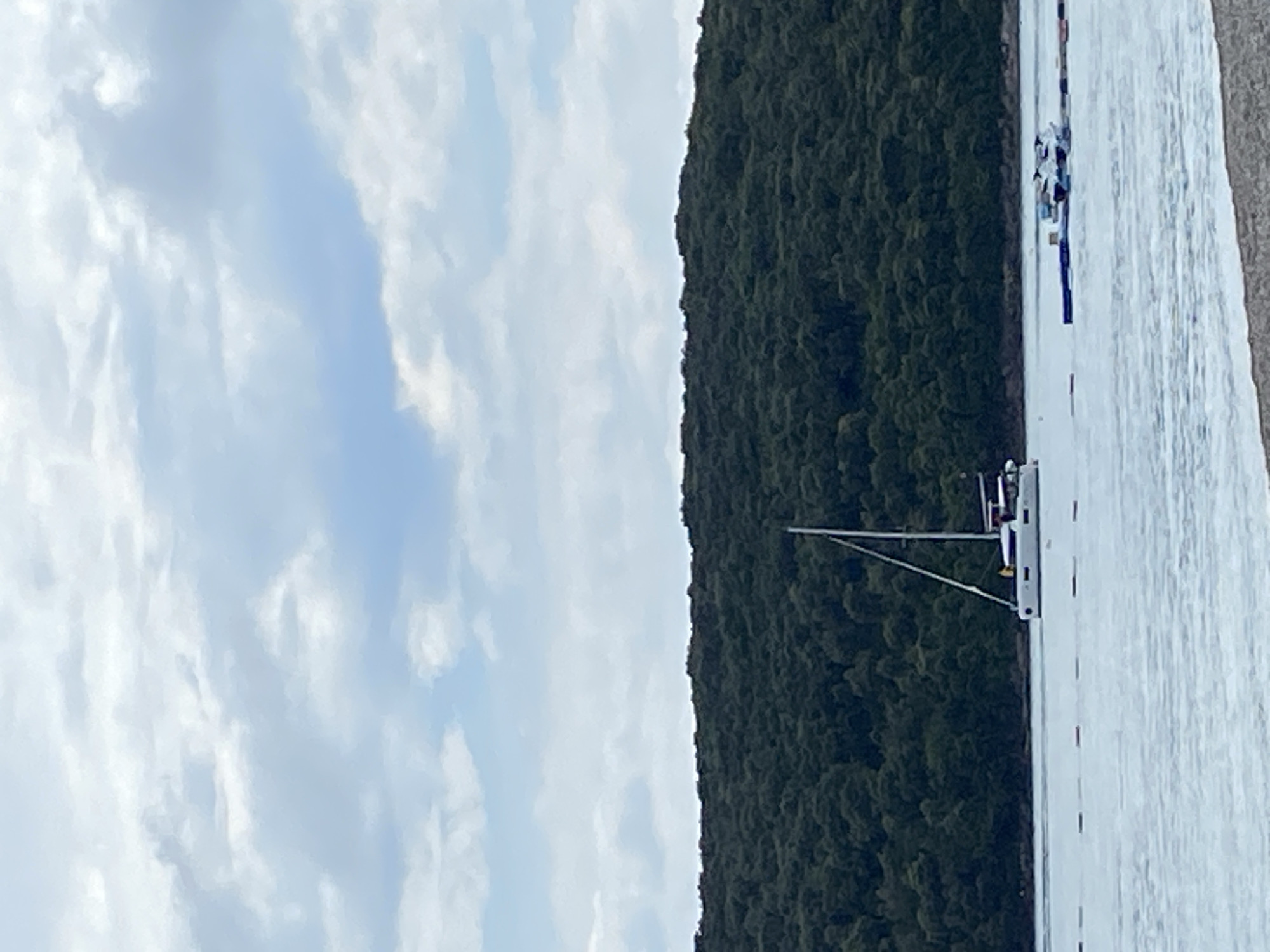 The image shows a landscape with a body of water, possibly a bay or lake, bordering a densely wooded area in Prijevor, Montenegro. The sky is partly cloudy. Several boats are visible on the water. One boat is more prominent, showing a mast and rigging. The location is likely a coastal or lakeside scene within the municipality of Tivat, Montenegro.