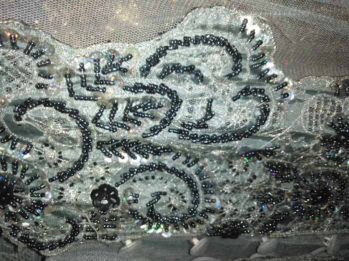 Intricate beading and sequin work adorn a light blue fabric. Dark, small beads trace ornate, swirling patterns. Clear sequins are scattered among the beads, adding a subtle shimmer. The fabric appears to be embellished, with a layer of sheer netting visible in the background. Overall, the image showcases detailed craftsmanship and a delicate aesthetic.