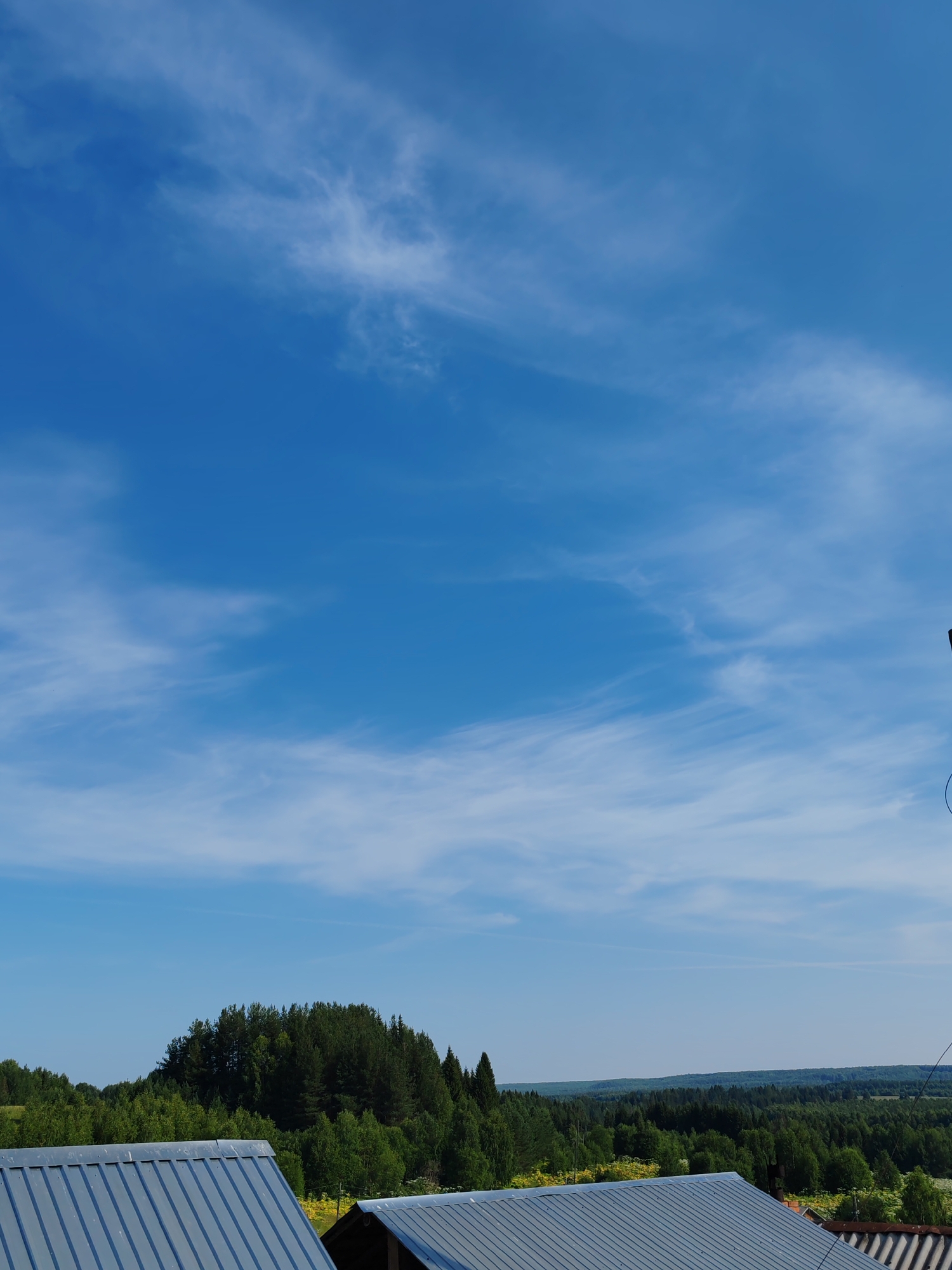 The image depicts a rural landscape under a bright blue sky with scattered wispy clouds. In the foreground, the metal roofs of two buildings are visible, one on the left and another partially on the right. Beyond the rooftops, a dense forest stretches across the middle ground, with a prominent tree-covered hill on the left. The background shows a hazy horizon of more distant forests. The scene appears to be taken from an elevated vantage point, possibly a rooftop or a higher elevation within the city of Chukhloma, Russia, given the rural and forested nature of the landscape. No people or animals are present in the image, and the overall atmosphere is peaceful and serene.