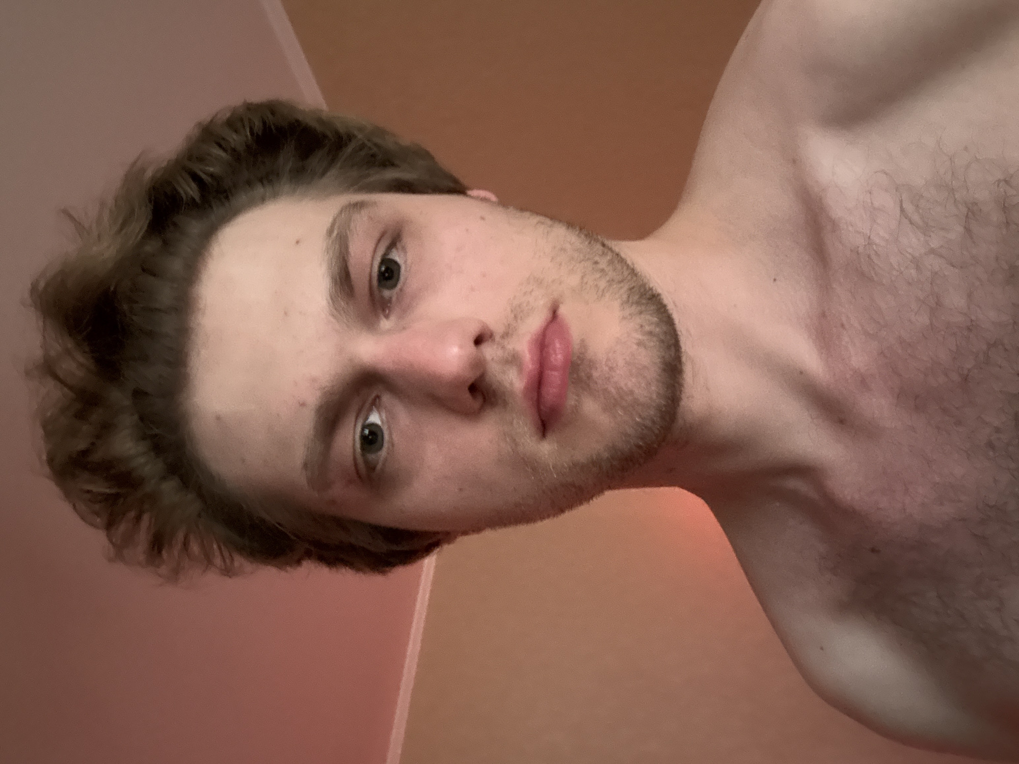 A close-up, head-and-shoulders selfie is captured from a frontal perspective. The subject, a young Caucasian male with light brown, tousled hair and a light beard, gazes directly at the camera. He is shirtless, revealing his chest and shoulders. The background consists of a wall with a subtle gradient of warm tones, transitioning from a peachy hue on the left to a muted orange on the right, suggesting artificial lighting. The environment appears to be an indoor setting.
