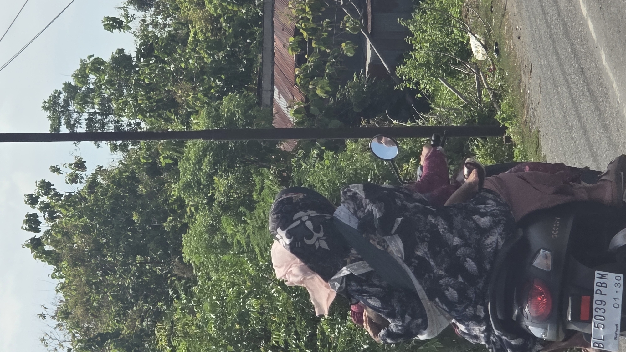 The image, taken in Teungoh Baro, Indonesia, shows two people on a black "Scoopy" scooter, located on an asphalt road. Lush green trees and vegetation line the background, with a building with a metal roof partially visible. A utility pole is situated on the left side of the image. The scooter's license plate reads "BL 5039 PBM" with an expiration date of "01.30". The individuals are dressed in clothing that covers their bodies, with the person in the front wearing a patterned scarf and the back passenger wearing a maroon outfit.