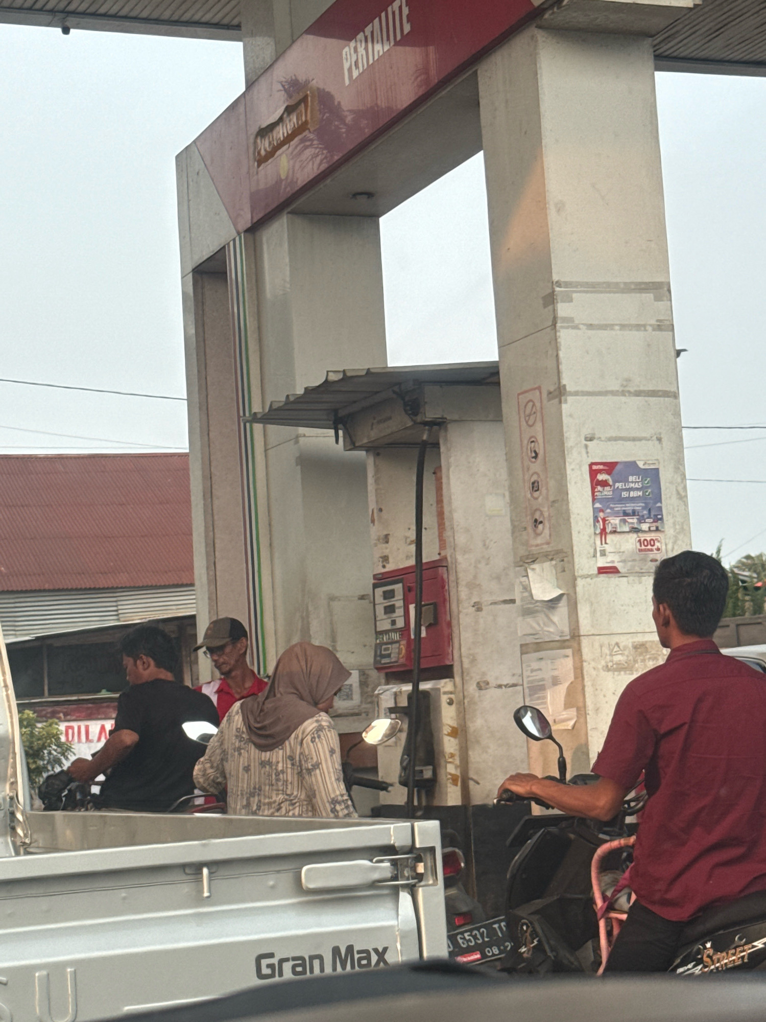 The image captures a scene at a Pertamina gas station in Mekar Mulya, Indonesia. In the foreground, a silver pickup truck with "Gran Max" branding is visible. Behind the truck, three individuals are present at a gas pump. A person in a black shirt is seated on a motorcycle, facing away from the camera. Another person, wearing a headscarf and a patterned shirt, is also on a motorcycle, appearing to interact with the pump. A third individual, wearing a red shirt and a cap, stands between them, possibly a gas station attendant or another customer. The gas pump structure, with its red and white branding, is a prominent element. Posters and signage are affixed to the pump's pillars. The environment suggests an outdoor setting with a cloudy sky.