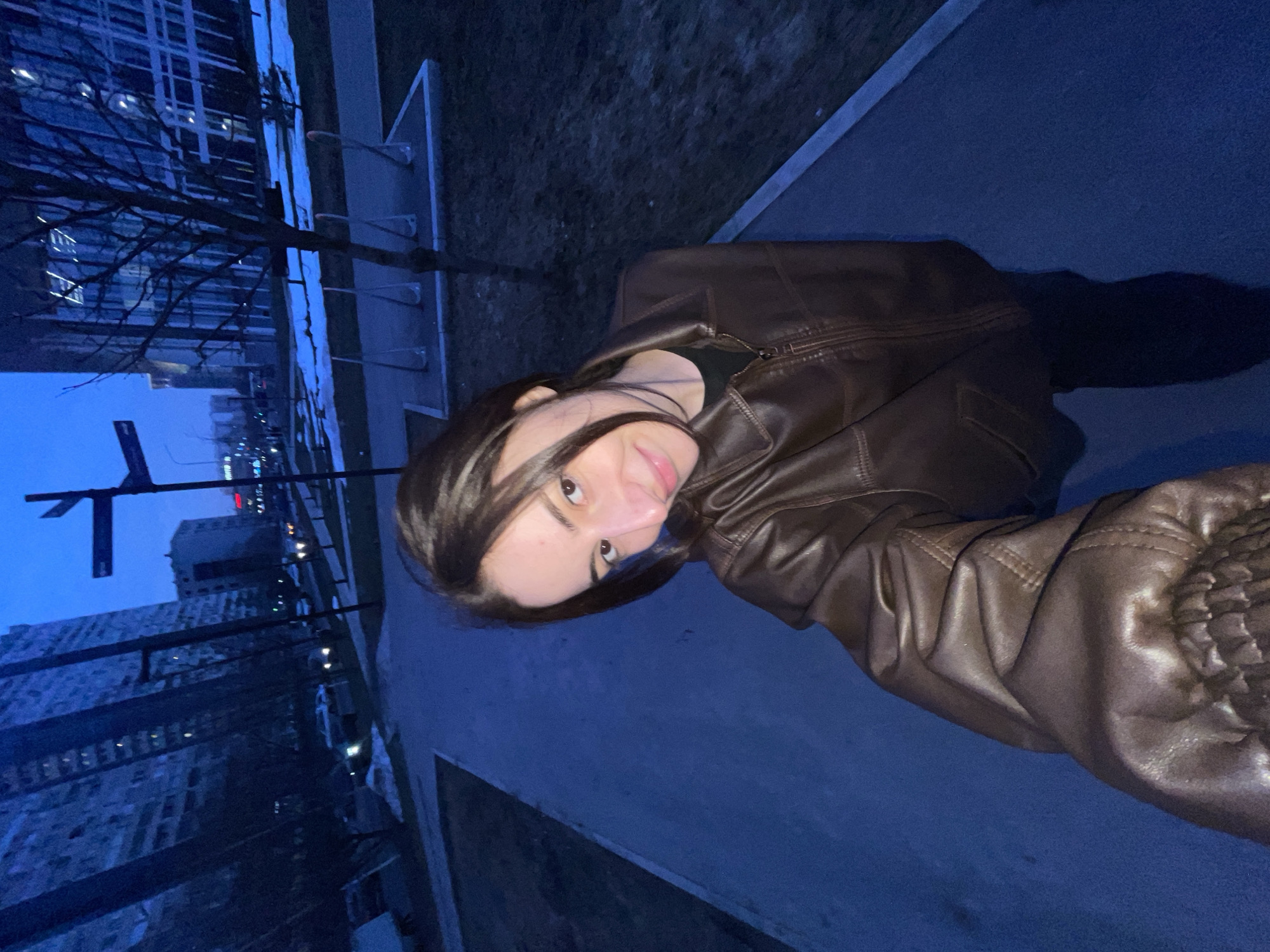 A young adult female, with dark hair parted on the side, wearing a dark brown leather jacket over a black top, is depicted in a selfie on a paved walkway in Moscow, Russia, during twilight hours. She holds the camera with her right hand, which is mostly out of frame, and is positioned slightly to the right of the central axis, looking directly at the viewer with a faint smile.
The environment indicates an urban district of Moscow. To the subject's immediate right, a dark ground area, possibly grass or dirt, contains patches of residual snow. A bare deciduous tree stands further back on the right, in front of a white concrete pad with several silver-colored bicycle racks. Behind these features, a modern building with a reflective glass facade is visible.
In the midground and background, the scene reveals a blend of architecture: multiple tall, multi-story buildings, characteristic of both residential and commercial high-rises found in Moscow. Some buildings display illuminated windows. Vehicle headlights and streetlights are visible on what appears to be a thoroughfare in the distance. A street sign on a pole is identifiable to the left of the bare tree. The sky is a deep blue, indicative of dusk or pre-dawn conditions.