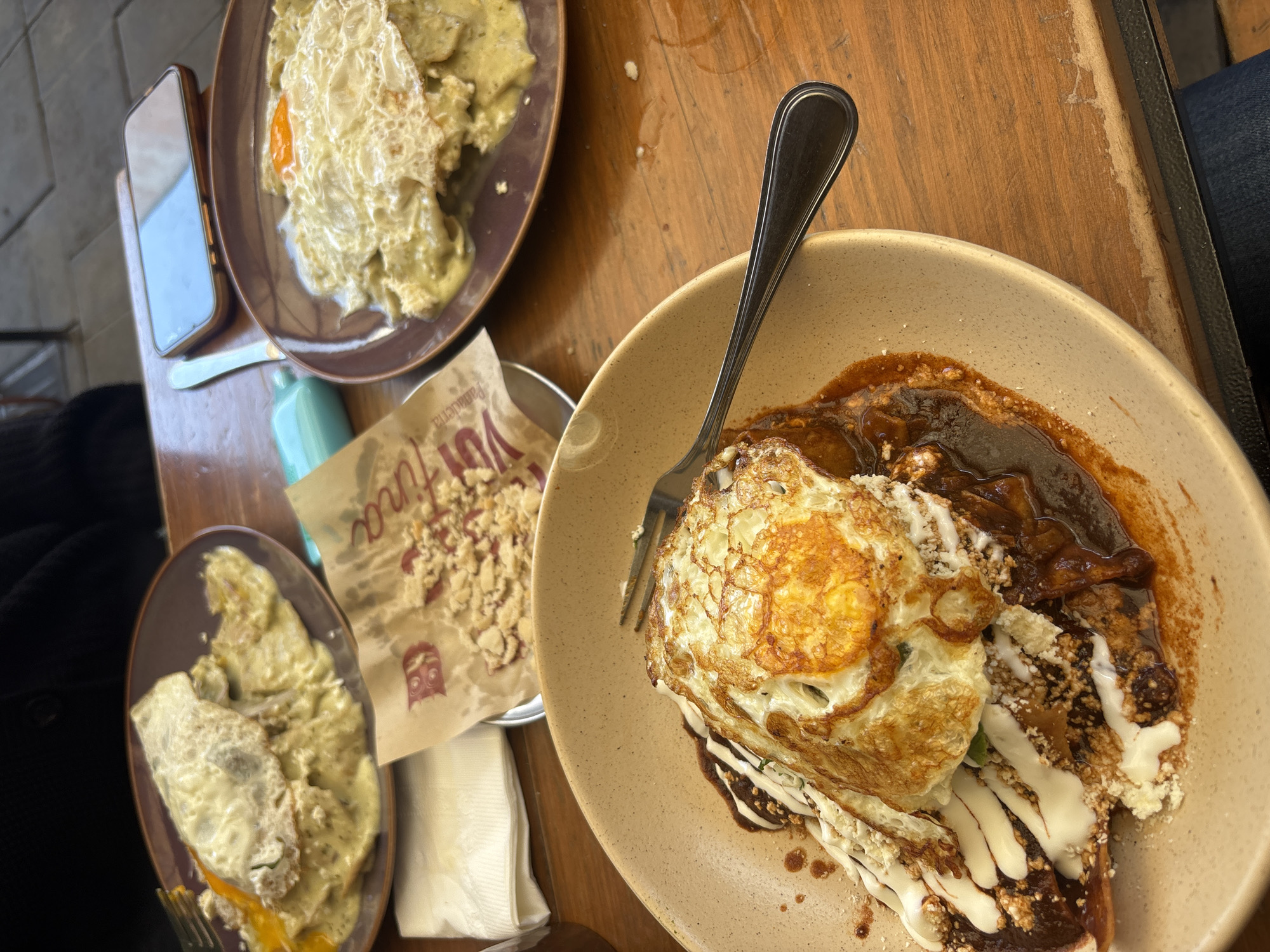 The image shows a restaurant table in San Andrés Cholula, Mexico, set with several plates of food. The table is made of wood.
Plate 1 (top left): Contains food covered in a greenish sauce, topped with a fried egg. The plate is dark brown.
Plate 2 (bottom left): Contains food covered in a yellowish sauce, also topped with a fried egg. The plate is also dark brown.
Plate 3 (right): Contains food covered in a dark brown sauce, topped with a fried egg and white sauce. A fork is placed on the plate. The plate is light brown.
Between the plates, there is a paper bag with crumbs inside. There is also a white napkin and a blue object. A smartphone sits next to the first plate. A person wearing jeans is visible on the right of the frame.