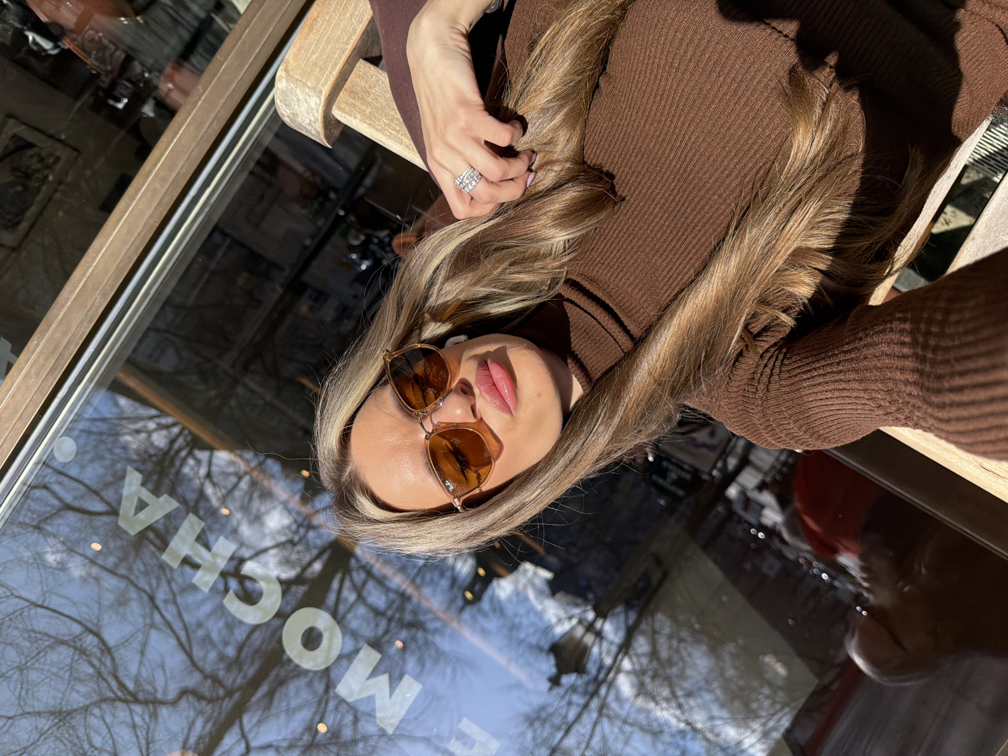 A woman with blonde hair and sunglasses is positioned near a window displaying the reflection of trees and the word "MOCHA." She is wearing a brown ribbed turtleneck sweater and a ring on her left hand. The scene is illuminated by natural light. There is no definitive evidence to pinpoint the specific location in Berlin, Germany. The presence of the word "MOCHA" suggests it could be a cafe or establishment offering mocha-based beverages.