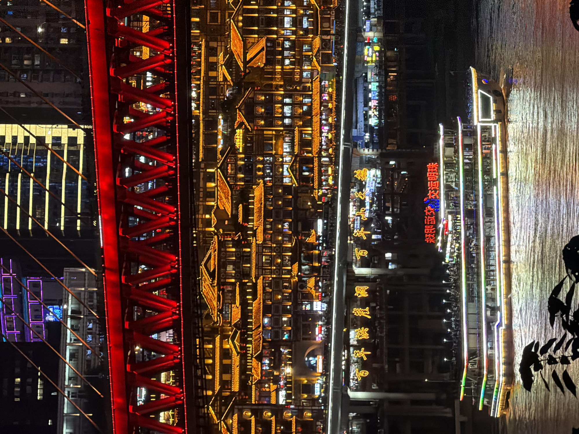 This is a nighttime, eye-level shot of a waterfront cityscape in Jiangbeicheng Subdistrict, China. In the foreground, dark foliage obscures the bottom of the frame, with the dark, rippling surface of a river occupying the lower half. A brightly lit, multi-decked passenger boat, decorated with colorful LED lights, is navigating the water. The boat has a visible hull and several illuminated windows. Behind the boat, a concrete structure resembling a bridge or elevated walkway spans the scene. This structure is adorned with large, bright Chinese characters in yellow. Above this, a dense cluster of traditional-style buildings, illuminated with warm yellow lights and featuring distinctive tiled roofs, cascades down the hillside. Even higher, a vibrant red, lattice-work bridge is prominently featured, illuminated with red lights, its structure crisscrossing the frame. In the background, modern high-rise buildings rise, some featuring colorful neon lighting, with windows casting yellow and white glows. The overall scene is one of a bustling, illuminated urban environment at night.