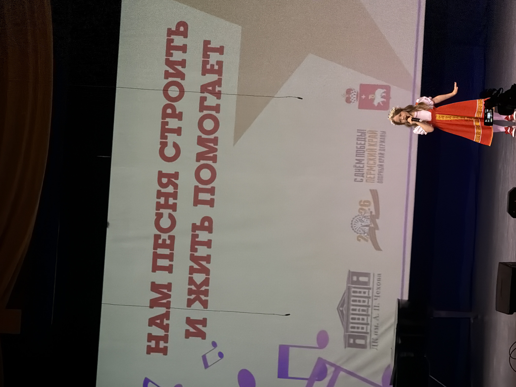 A young girl in traditional Russian attire sings on a stage in front of a screen. The screen displays a banner in Russian that reads "Our song helps build and live" in red letters, followed by music notes. Above this, the banner also includes logos for "DK im. A. P. Chekova" and "Permsky Krai", indicating the location is Perm, Russia. The stage has a dark backdrop. The screen also has the text "С Днём Победы Пермский Край" (Happy Victory Day Perm Krai), which makes it likely that the event is related to the Russian holiday Victory Day, which is celebrated on May 9th.