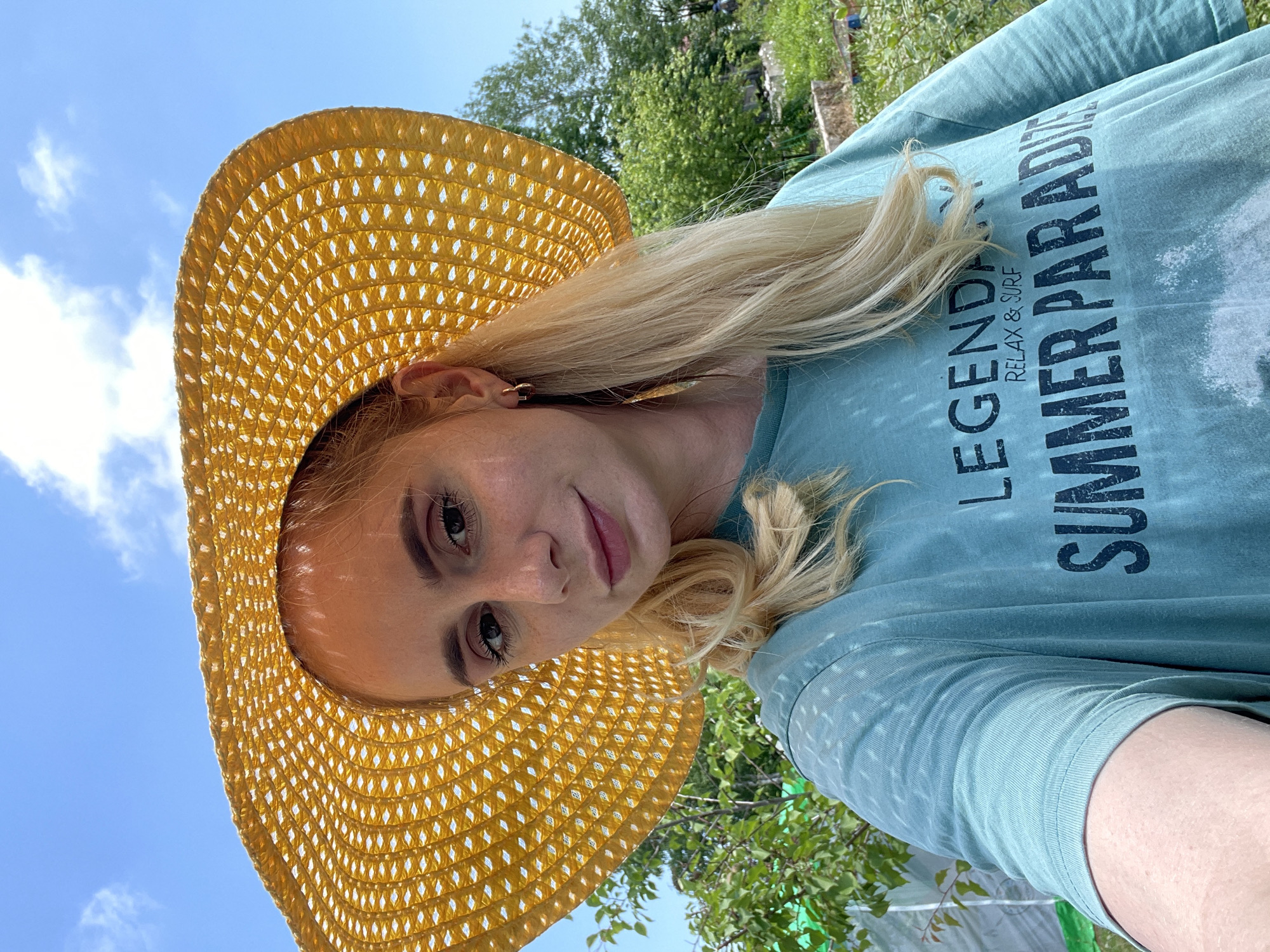 A young woman with blonde hair and fair skin is pictured in a close-up shot, wearing a large, yellow, woven sun hat and a light blue t-shirt. The t-shirt has text printed on it, with the words "LEGENDARY RELAX & SURF SUMMER PARADISE" visible. She has light makeup on and is wearing a small hoop earring in her visible ear. The background suggests an outdoor setting with lush green trees and foliage, under a clear blue sky with scattered clouds. The overall scene appears to be taken in a natural or garden environment. There is no indication of the location being the city of Knevichi, Russia.