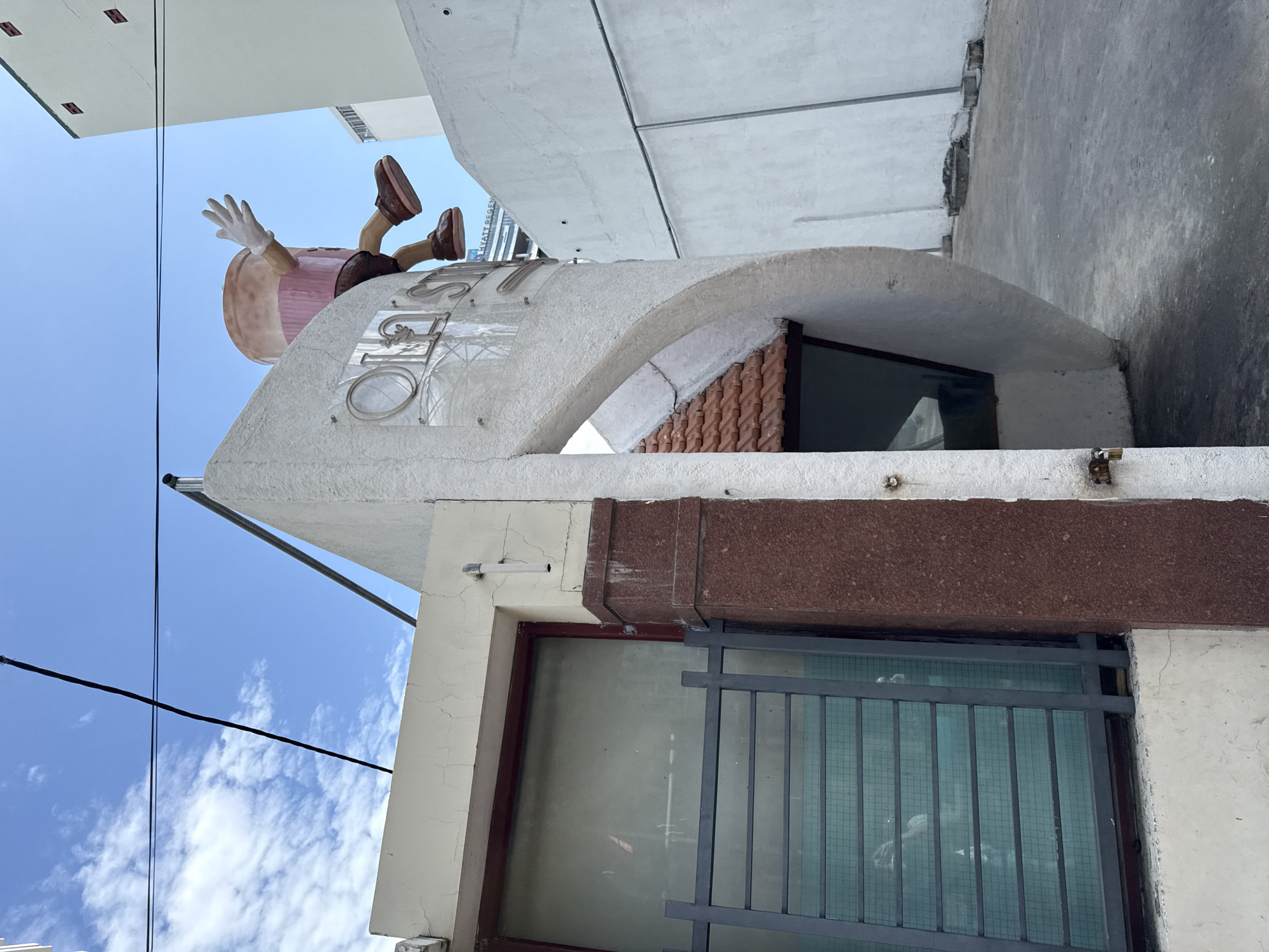In Nha Trang, Vietnam, an outdoor scene shows a whimsical giant ice cream cone sculpture with arms and legs waving from atop a white, curved building. Below the sculpture, a dark red, granite-clad pillar is visible on the left, next to a window covered by a grey metal grille. A tiled, brown roof extends from the building, creating an overhang. Power lines crisscross the bright blue sky, dotted with white clouds. The background features a white building and a glimpse of a modern skyscraper.