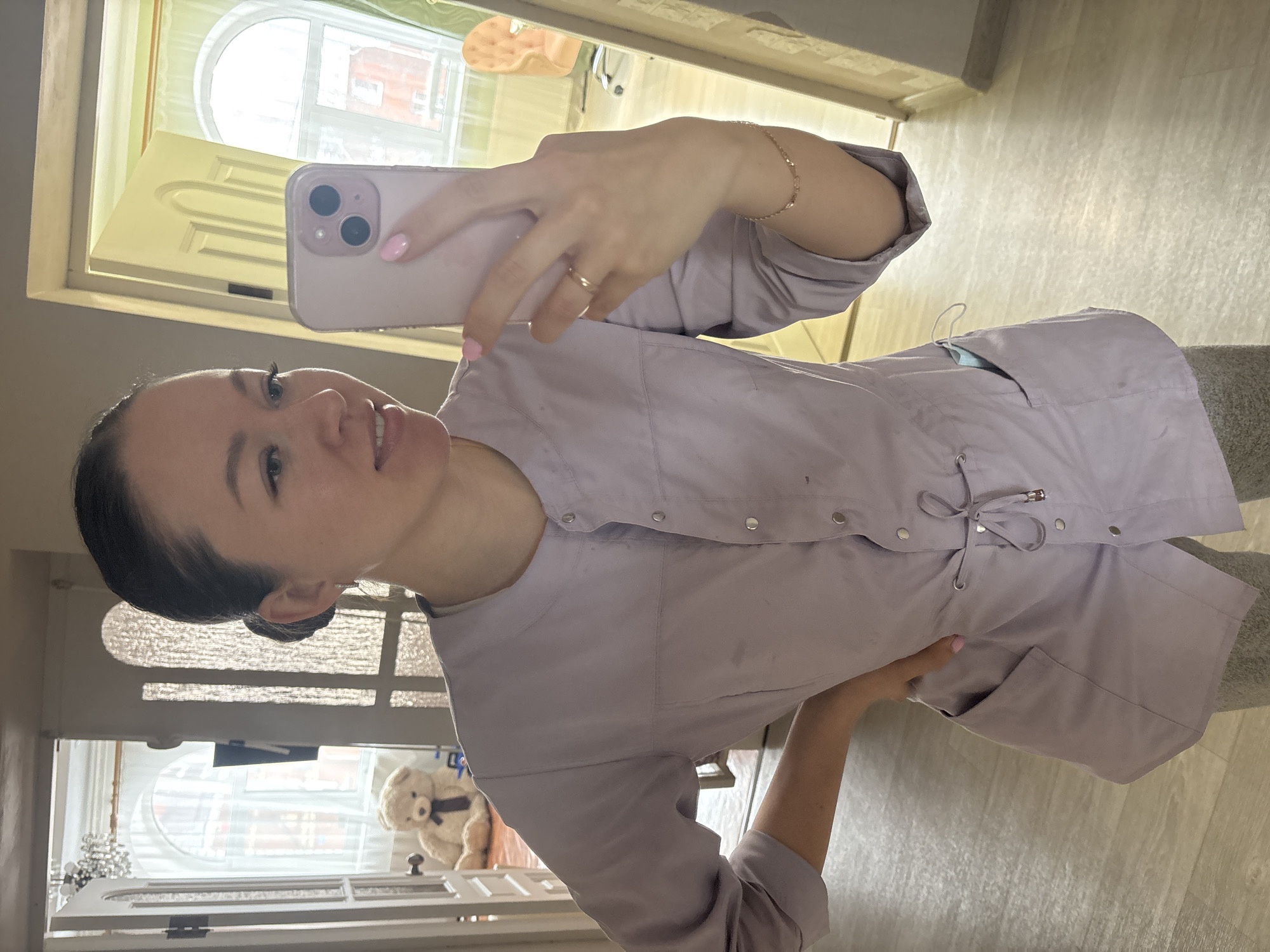 A woman is taking a selfie indoors. She is wearing a light purple button-down shirt and holding a pink smartphone. A window with decorative grilles and a teddy bear on a shelf is visible in the background. The location is suggestive of an interior space, possibly in Ferma, Russia based on the tags.