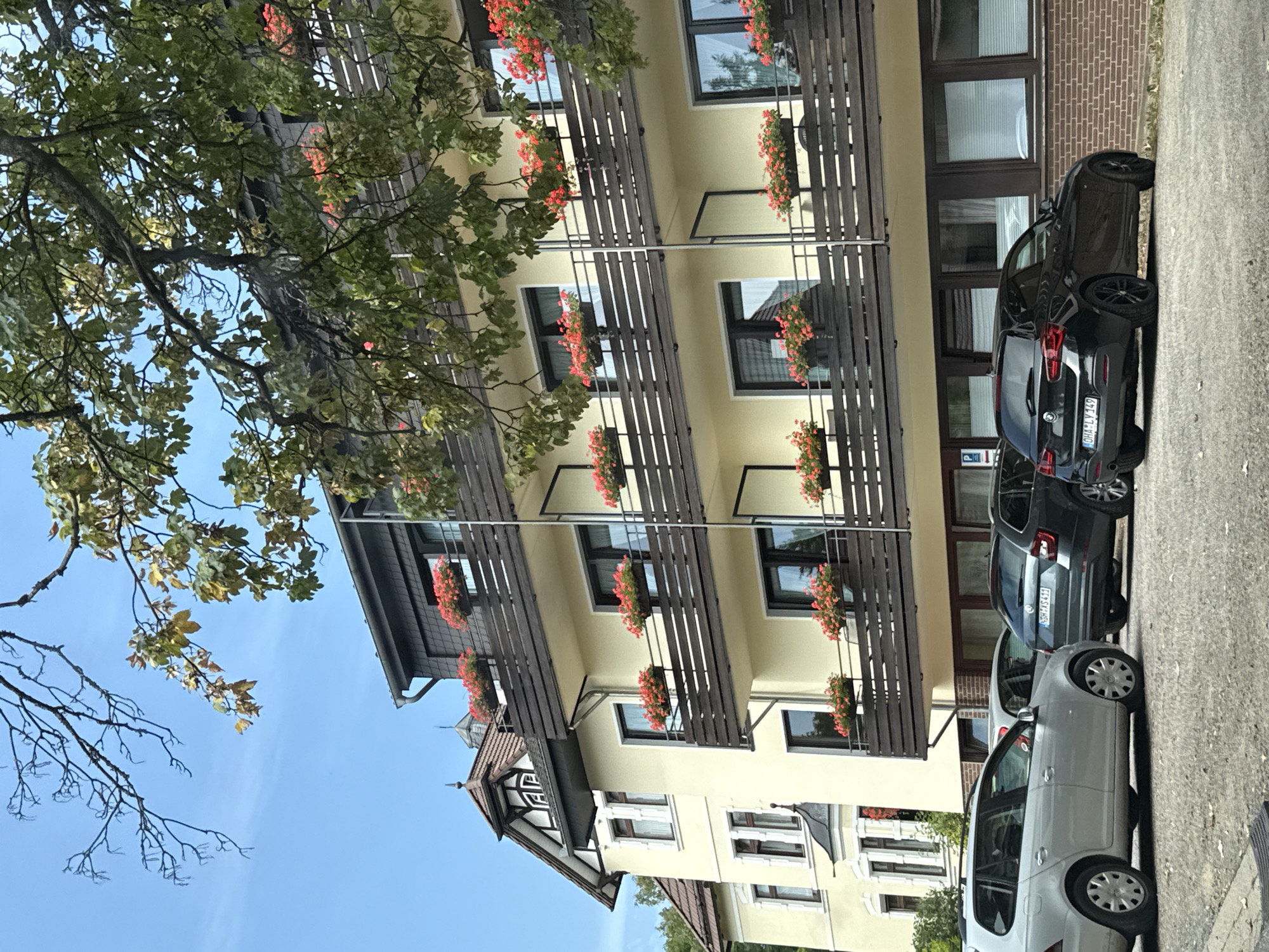 The image depicts a multi-story building with a light yellow facade, featuring several balconies adorned with red flowers in planters. The building is situated in Bad Sachsa, Germany, as indicated by the license plate of one of the vehicles. In front of the building, a row of cars is parked on a paved surface. Among them is a silver station wagon on the left, a dark gray station wagon in the middle with the license plate "NDH-XS411," and a black BMW hatchback on the right with the license plate "OHA-LY 149." The scene is observed from a slightly elevated perspective, with the branches of a tree, partially covered in leaves, extending into the frame from the top. The sky is clear and blue.