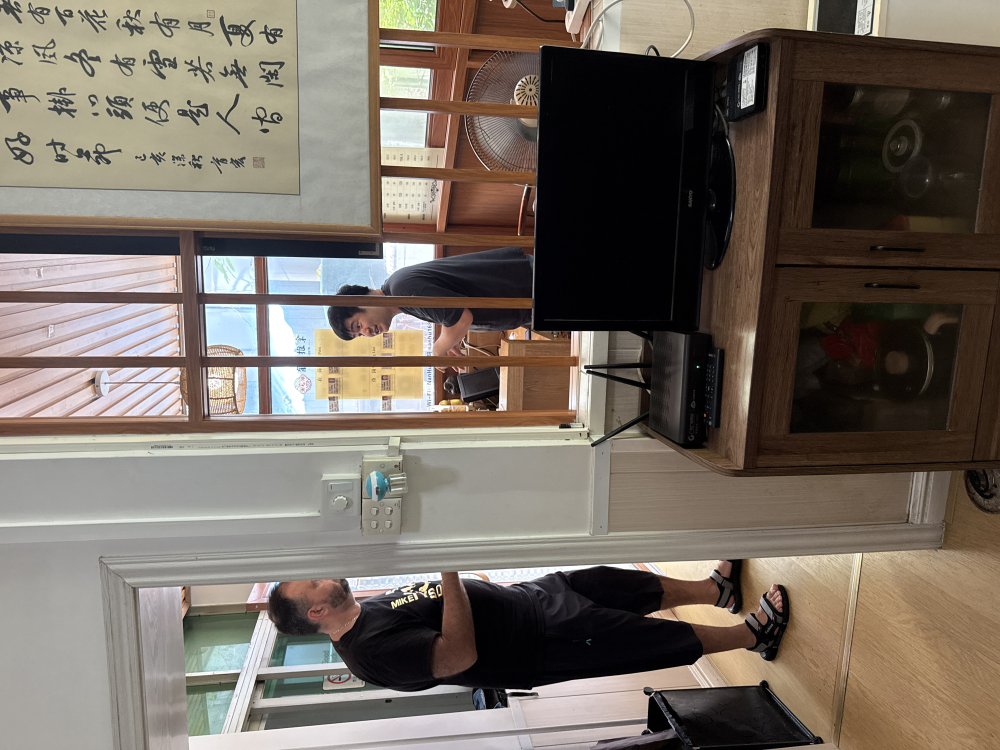 This image depicts the interior of what appears to be a restaurant or shop in Huizhou, China. In the foreground, a man wearing a black t-shirt and black shorts stands in a doorway, facing away from the camera. He has a gold chain around his neck and is wearing sandals. To his right, through a windowed partition, another man in a dark t-shirt is visible, talking and gesturing. He is standing behind a wooden counter. On a wooden cabinet in the foreground, a black television screen sits atop a router and a set-top box. Behind the partition, a ceiling fan is visible, and traditional Chinese calligraphy scrolls are displayed on the wall. The environment suggests a casual establishment, possibly a local eatery or a small business.