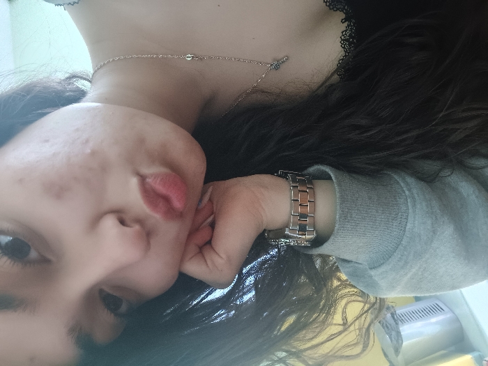 The image displays a close-up, angled indoor portrait of a female individual in Gorna Oryahovitsa, Bulgaria. The subject is a young woman with long, dark, wavy hair partially obscuring her face and left shoulder. She is positioned with her head tilted, looking towards the camera, and her lips are pursed in a reddish-pink pout. Her right hand (from the viewer's perspective) rests against her right cheek and jawline. Visible blemishes are present on her right cheek. She wears a grey long-sleeved garment, with a black top featuring lace trim visible beneath it on her left side. A silver-colored metal link watch adorns her right wrist, and a thin silver-colored chain necklace with a small cross pendant is visible on her chest. The background consists of a plain, light-colored wall, with a light-colored plastic container or bin with vertical slats partially visible in the lower-left corner, next to a yellow surface on the far left. No other individuals or overt interactions are present.