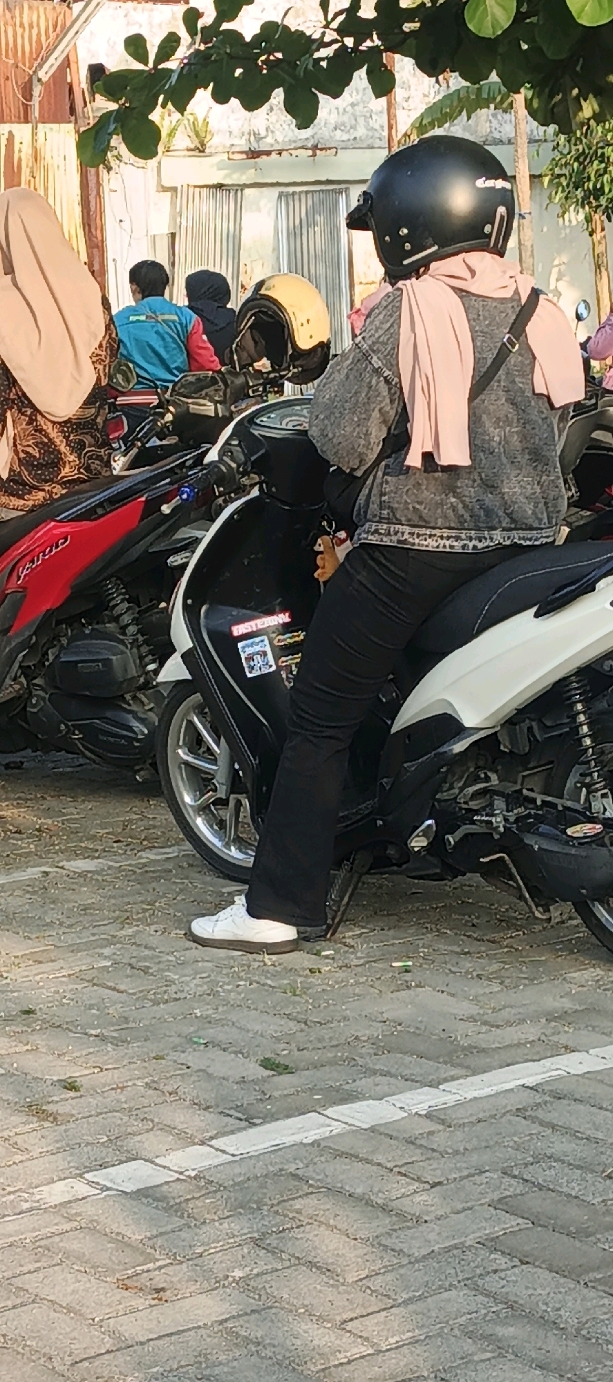 The image depicts a street scene in Salatiga, Indonesia. The primary focus is on a person seated on a white and black motorcycle. This individual is wearing a black helmet, a light pink hijab, a grey denim jacket, and black pants. Their left foot, clad in a white sneaker, is resting on the ground. To the left of this motorcycle, a red motorcycle is partially visible. In the background, two other individuals are seated on a motorcycle, one wearing a blue jacket and the other a dark hijab. The ground is paved with interlocking grey bricks. Green foliage from trees overhangs the scene from the top. The overall atmosphere suggests a casual outdoor setting, possibly a parking area.