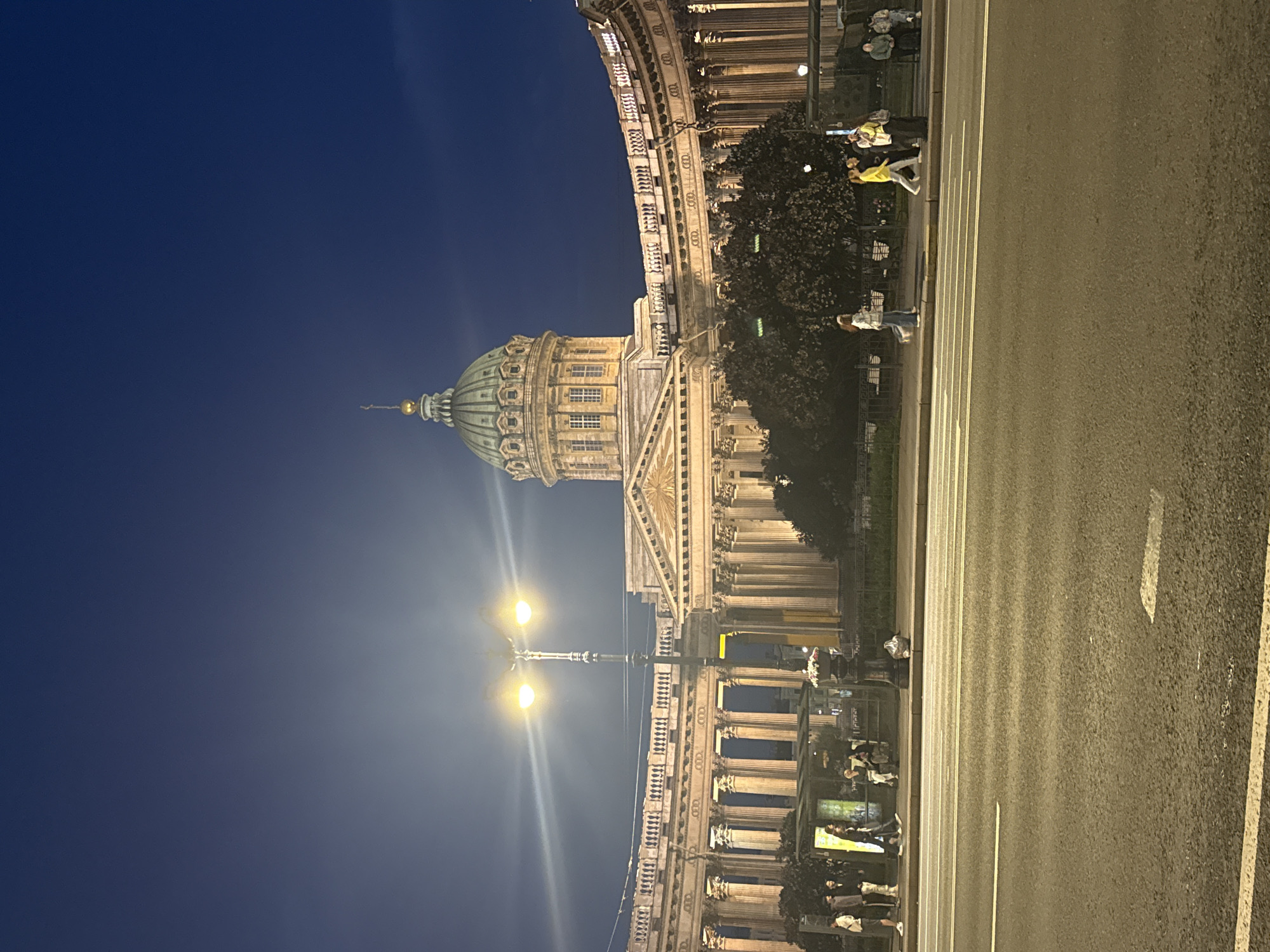 This image shows a wide, paved road in front of the Kazan Cathedral in Saint Petersburg, Russia, at dusk. The cathedral's grand Neoclassical facade, characterized by its semi-circular colonnade and central dome, is illuminated by artificial lights. Several people are visible walking and standing on the sidewalk near a bus stop, some illuminated by the streetlights. The sky is a deep blue, transitioning to lighter shades near the horizon. Streetlights cast a warm glow, and the road surface shows signs of wear, with visible lane markings. Trees are planted along the sidewalk, their leaves dark against the illuminated building.
