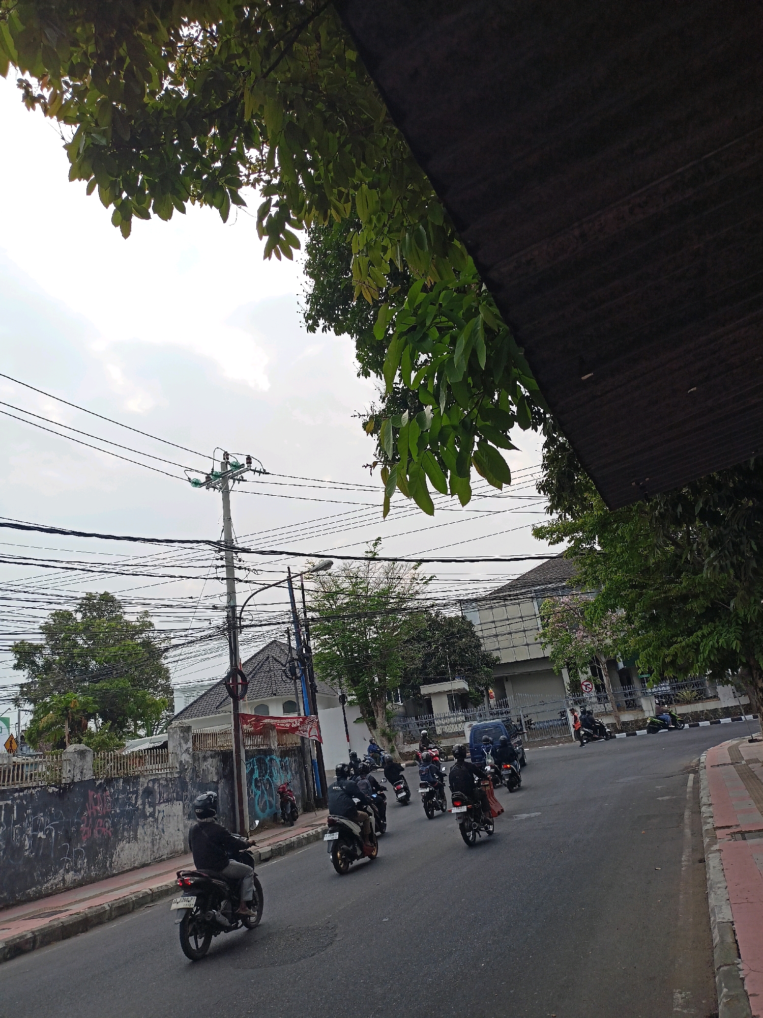 The image depicts a street scene in Magelang, Indonesia, on a cloudy day. The foreground is dominated by lush green foliage hanging from trees and a dark, textured overhead structure, possibly a roof or awning. Below, a paved road runs diagonally through the frame, with a sidewalk on the right side marked by red and gray concrete. A procession of motorcycles, each ridden by a person wearing a helmet, travels along the road. In the background, a utility pole with numerous tangled electrical wires is prominent, accompanied by buildings with tiled roofs and modern glass facades. Graffiti adorns a low wall on the left side of the road, adding a touch of urban texture. Several trees are scattered throughout the scene, some bearing pink flowers, and a blue van is visible amongst the motorcycles. The overall impression is of a busy street in a tropical urban environment.