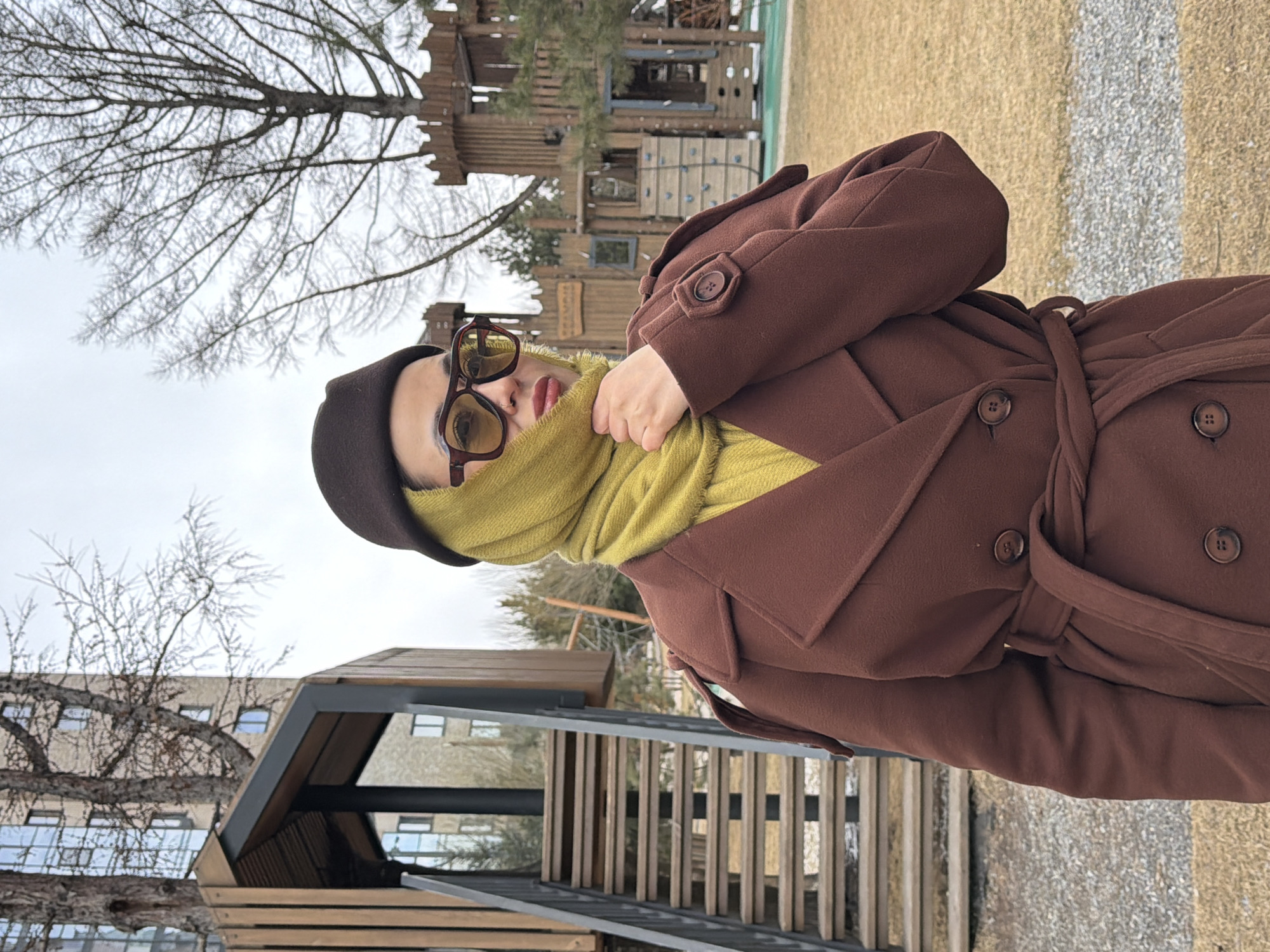 Here is a description of the image:
A person is seen wearing a brown coat, a dark cap, and sunglasses with a yellow scarf wrapped around their neck. The person appears to be outdoors in a public park or recreational area. A modern wooden structure, possibly a shelter or playground, is visible in the background, along with leafless trees indicating a cold season. In the far background, there is a building with numerous windows. The ground has a mix of gravel and grass.
Based on the background elements and the presence of a wooden playground structure, the location is possibly a public park or square. Since Krasnoyarsk is requested, it is likely that this picture was taken in Krasnoyarsk, Russia, although specific identification landmarks are absent from this image.
